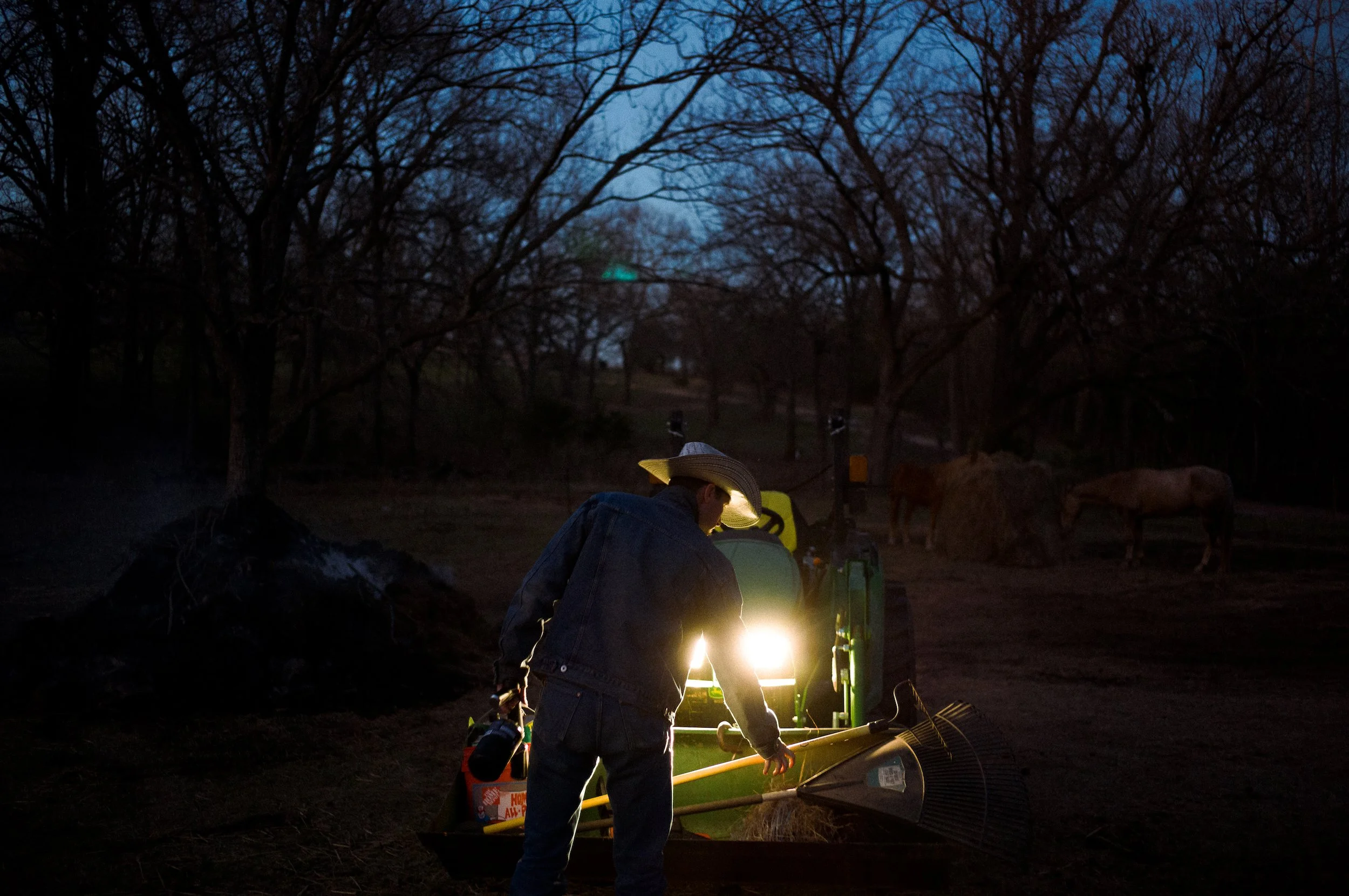 A person wearing a wide-brimmed hat and denim jacket working on equipment outdoors at night, illuminated by artificial light, with trees in the background.