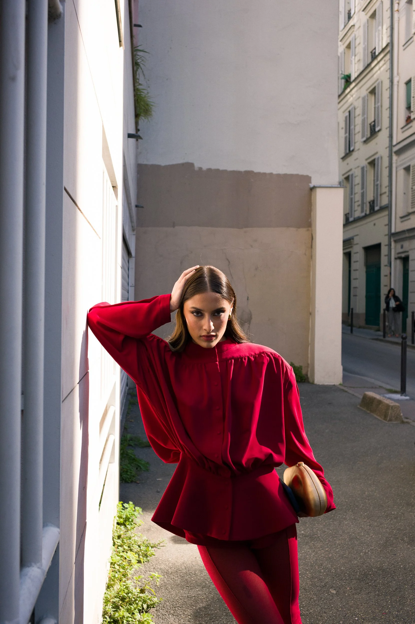 At Paris Fashion Week, A woman wearing a red outfit is standing outdoors in an alleyway with her hand on her head, holding a hat in her other hand, with buildings and a sidewalk in the background.