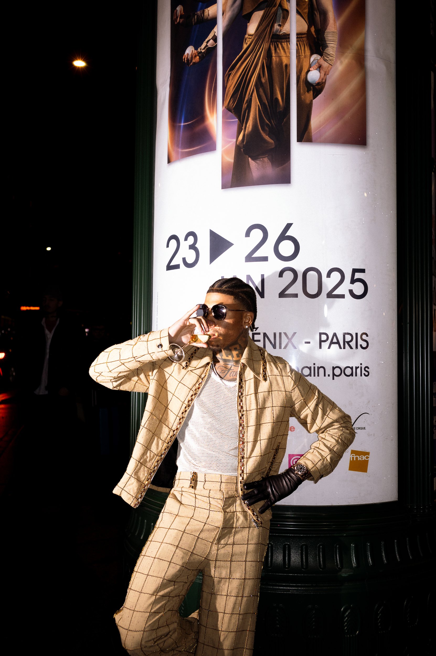 At Paris Fashion Week, A man with sunglasses, jewelry, a beige checked suit, and a white shirt poses with one hand on his hip and the other adjusting his sunglasses in front of a large advertisement poster on a city street at night.