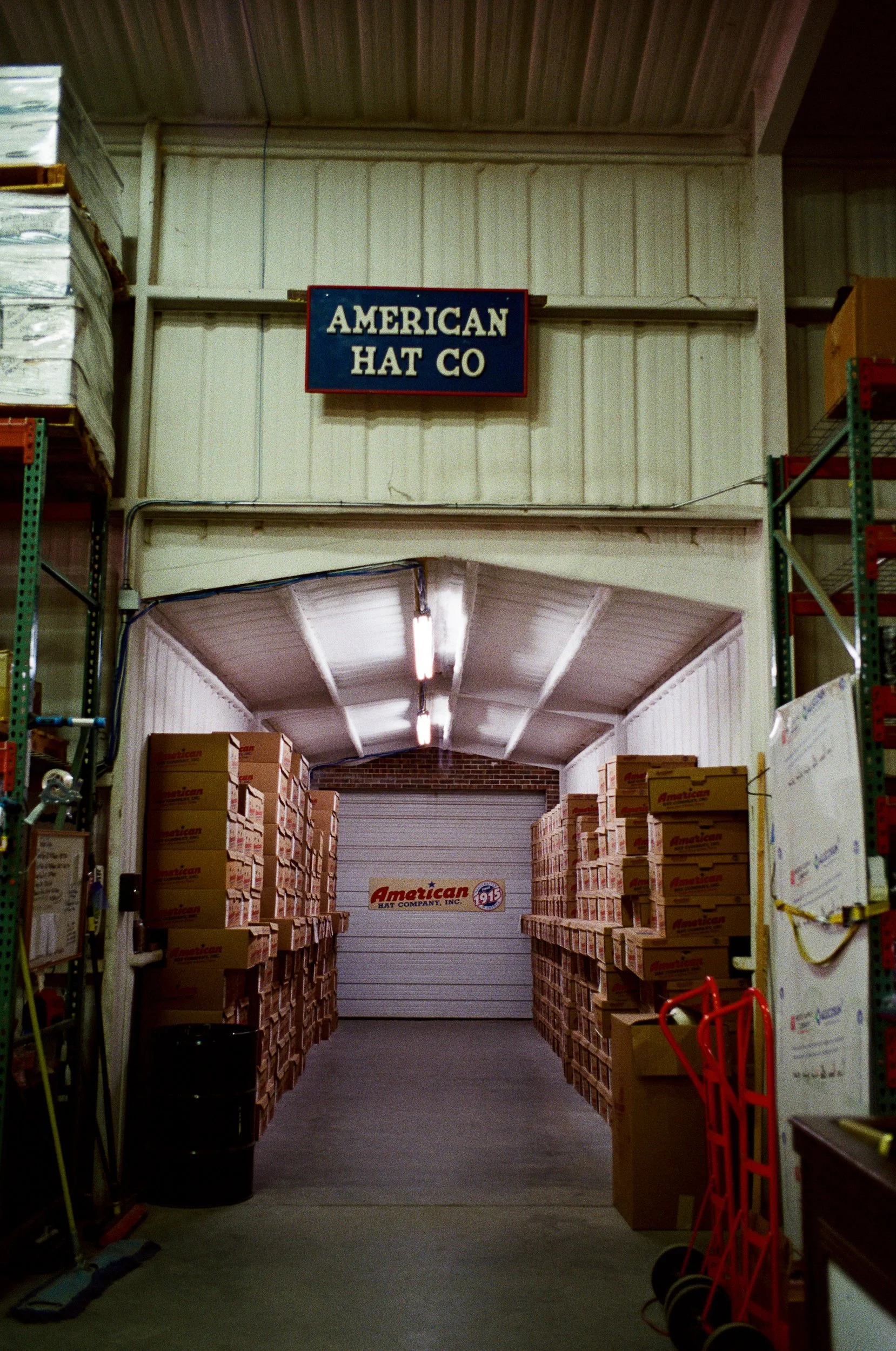 A warehouse aisle with shelves full of stacked boxes labeled "American Hat" and a garage-style door at the back. Sign reading "American Hat Co" hangs above the aisle.