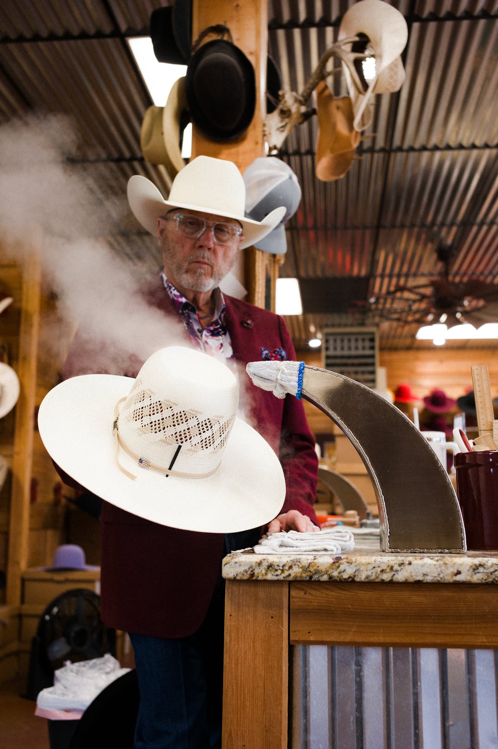 A man with glasses and a beard wearing a white cowboy hat, a burgundy blazer, and a colorful shirt, shaping a hat with steam. Hats hang on a wooden beam above him, and he appears to be looking at the camera.