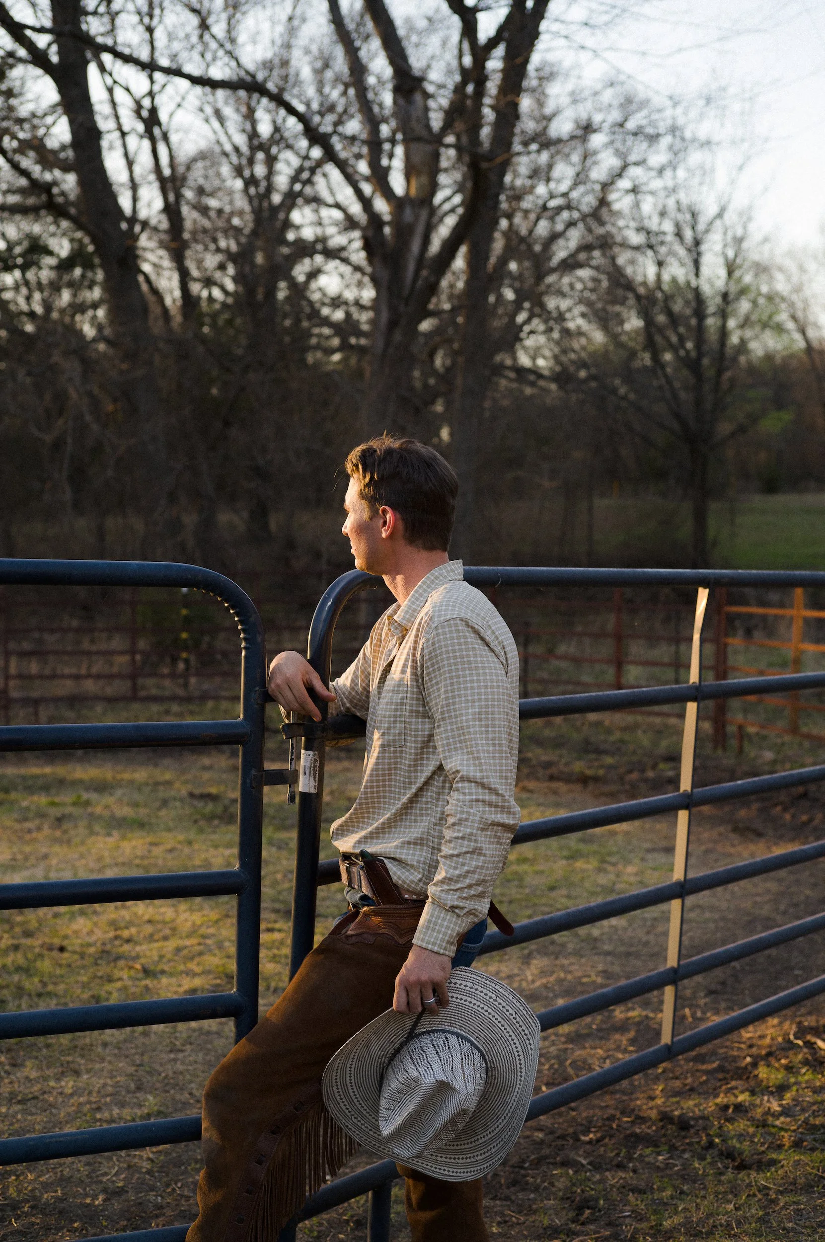 Cowboy wearing a plaid shirt and brown pants holding a wide-brimmed hat and a cowboy hat, standing outdoors near a metal fence with trees in the background during sunset.