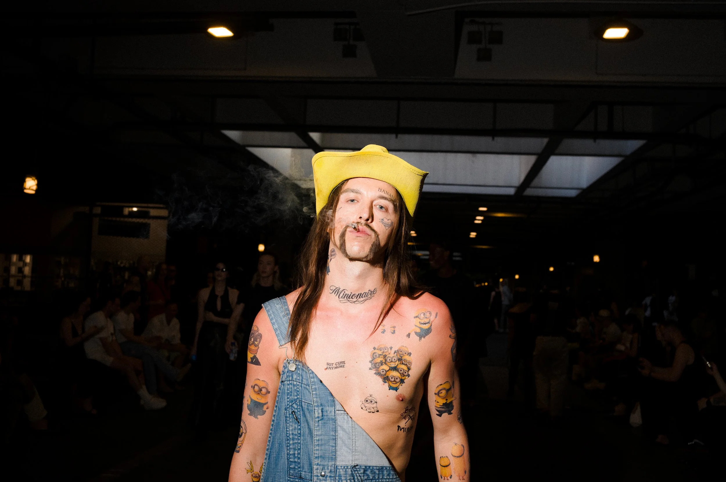 A person with long hair, tattoos, and facial hair, wearing a yellow beanie and a denim dress, standing indoors in front of a group of people.