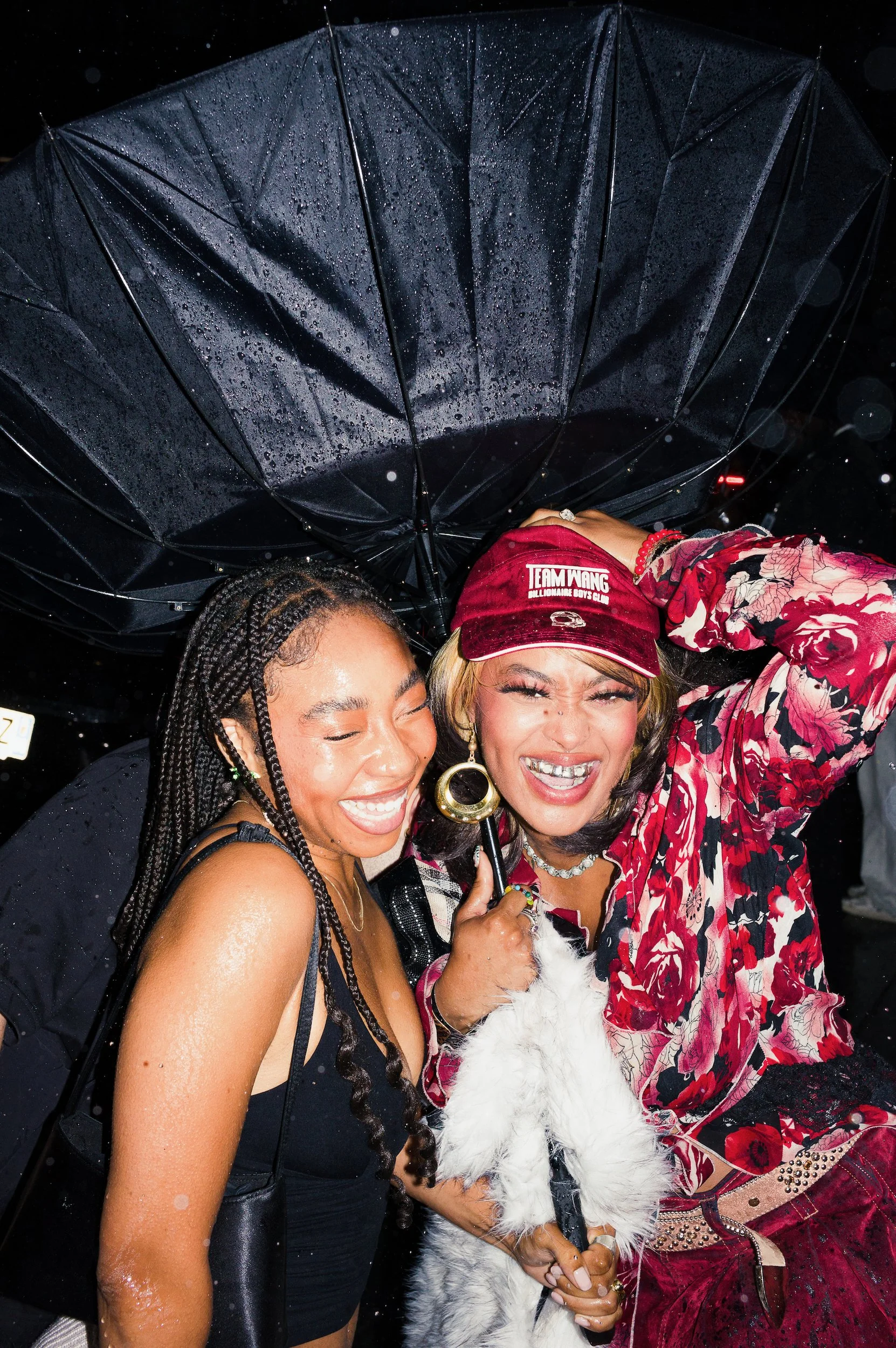  At Paris Fashion Week, two women smiling and standing close together under a black umbrella with raindrops on it, at night.