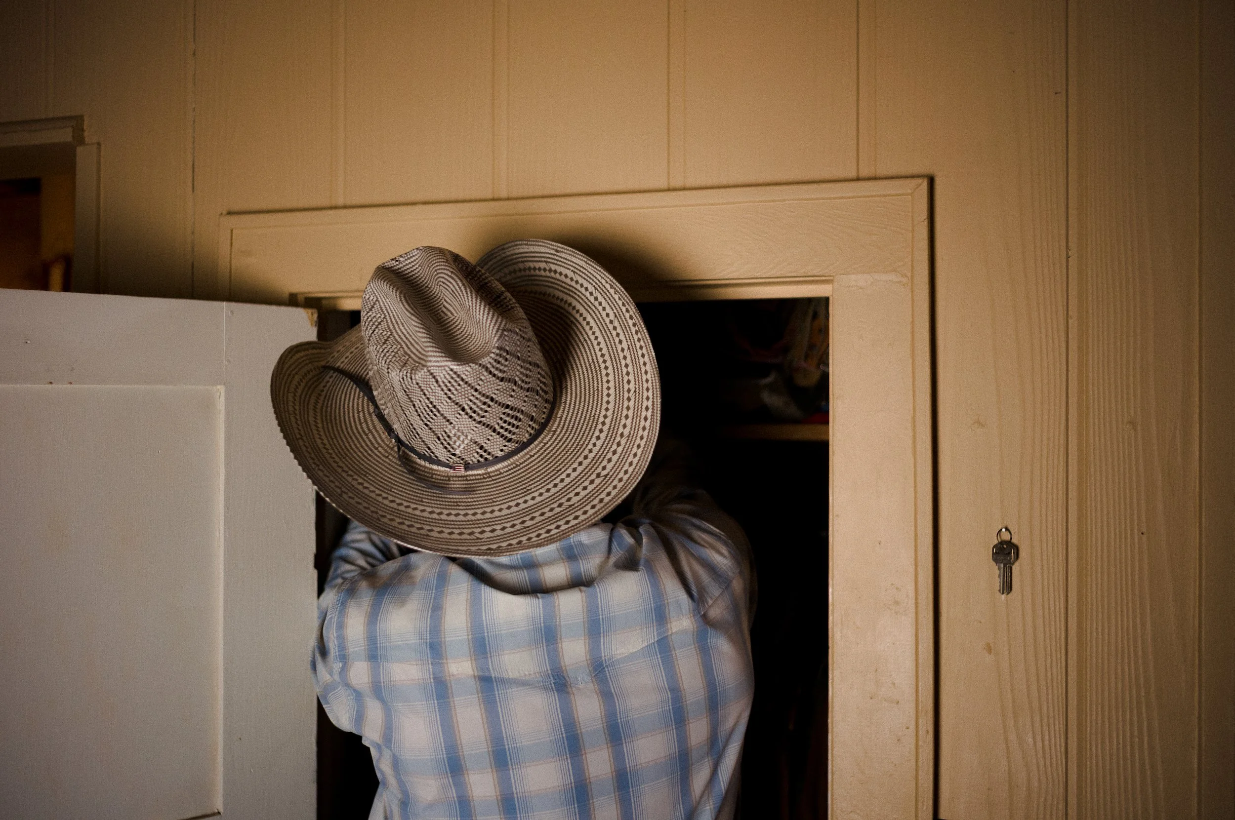 Cowboy wearing a checkered shirt and a wide-brimmed hat standing in front of a wooden door and a white wall; a set of keys hanging on the door handle.