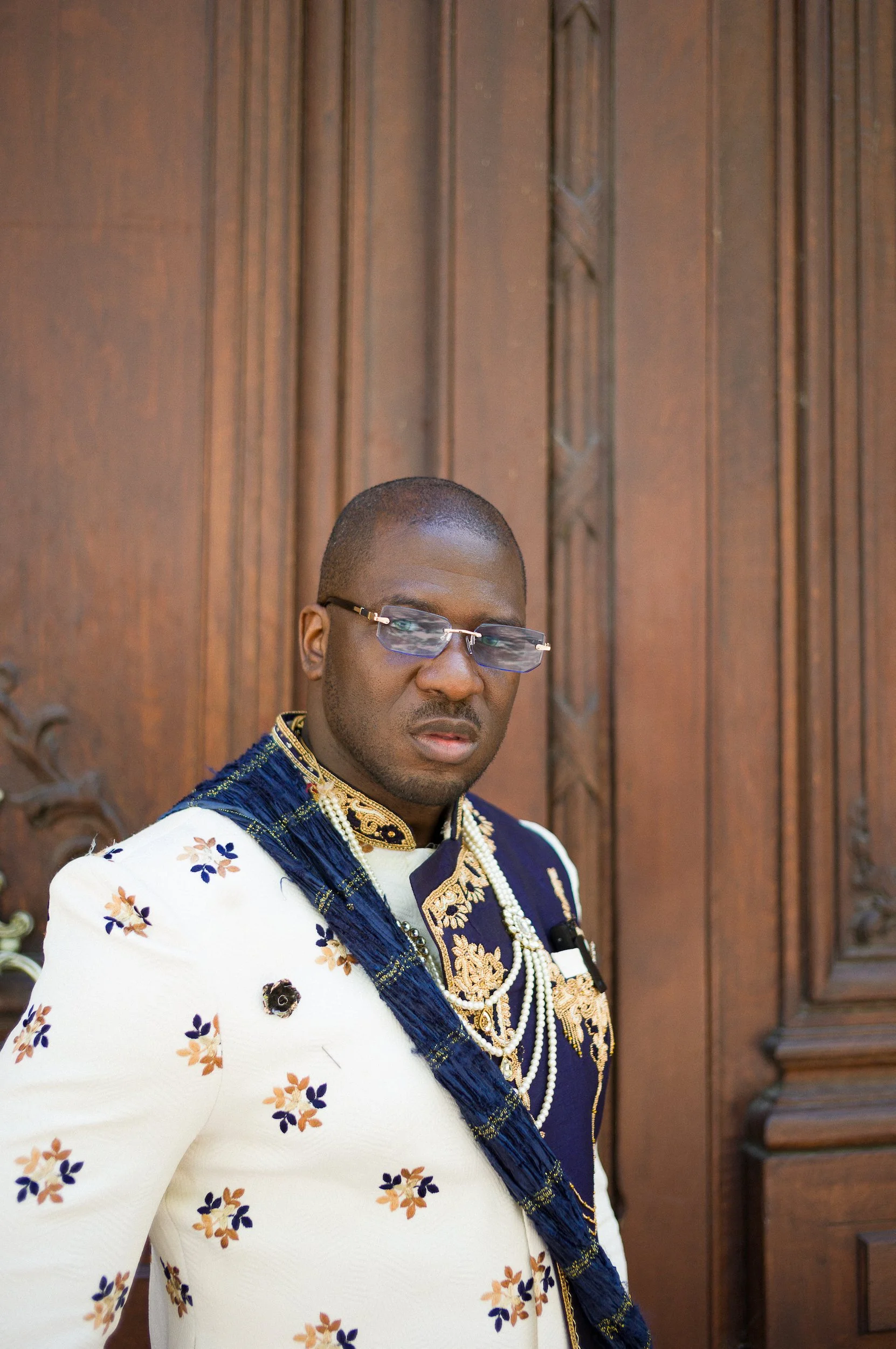 At Paris Fashion Week, A man dressed in traditional African attire standing in front of a wooden door, wearing glasses, and decorated with gold jewelry and a beaded necklace.