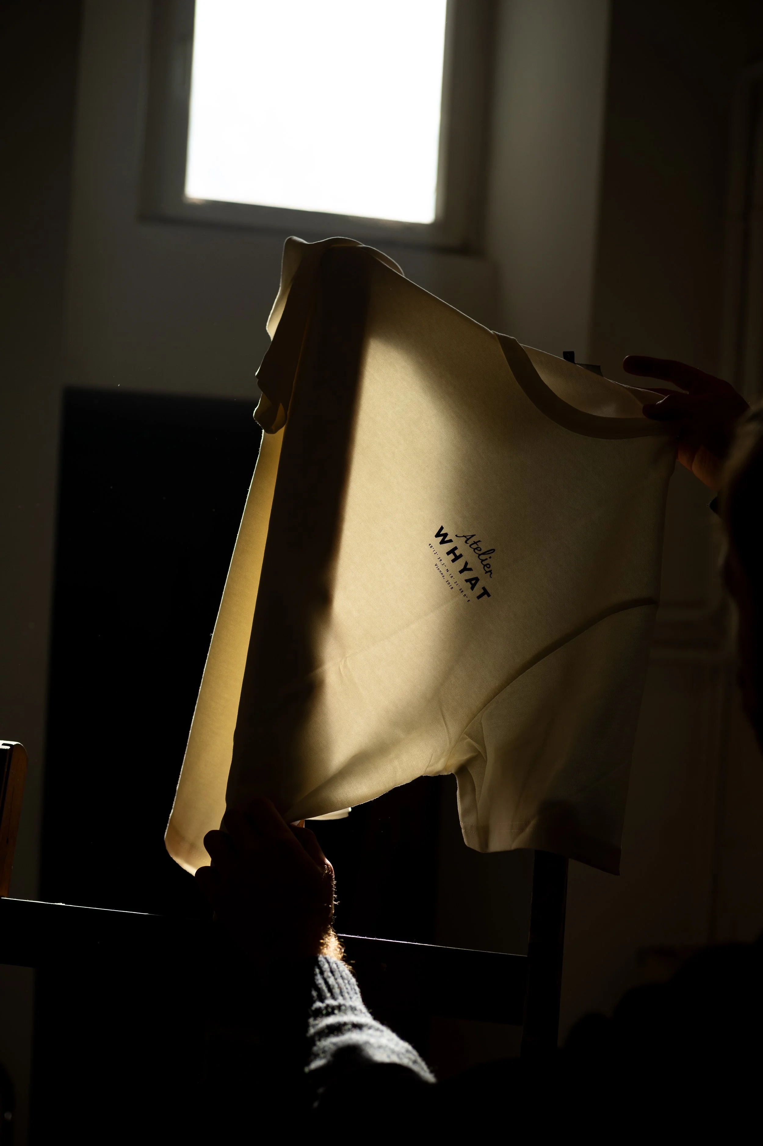 Person holding a canvas or bag with the text "Atelier WHYAT" printed on it, backlit with sunlight coming through a window.