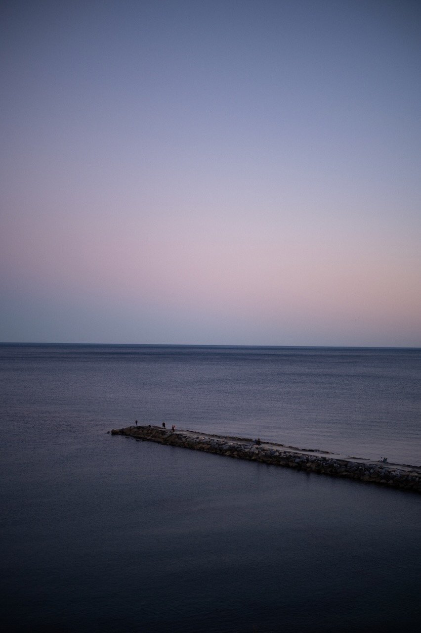 In Spain, A calm ocean with a small breakwater extending into the water, under a pastel-colored sky at dusk or dawn.