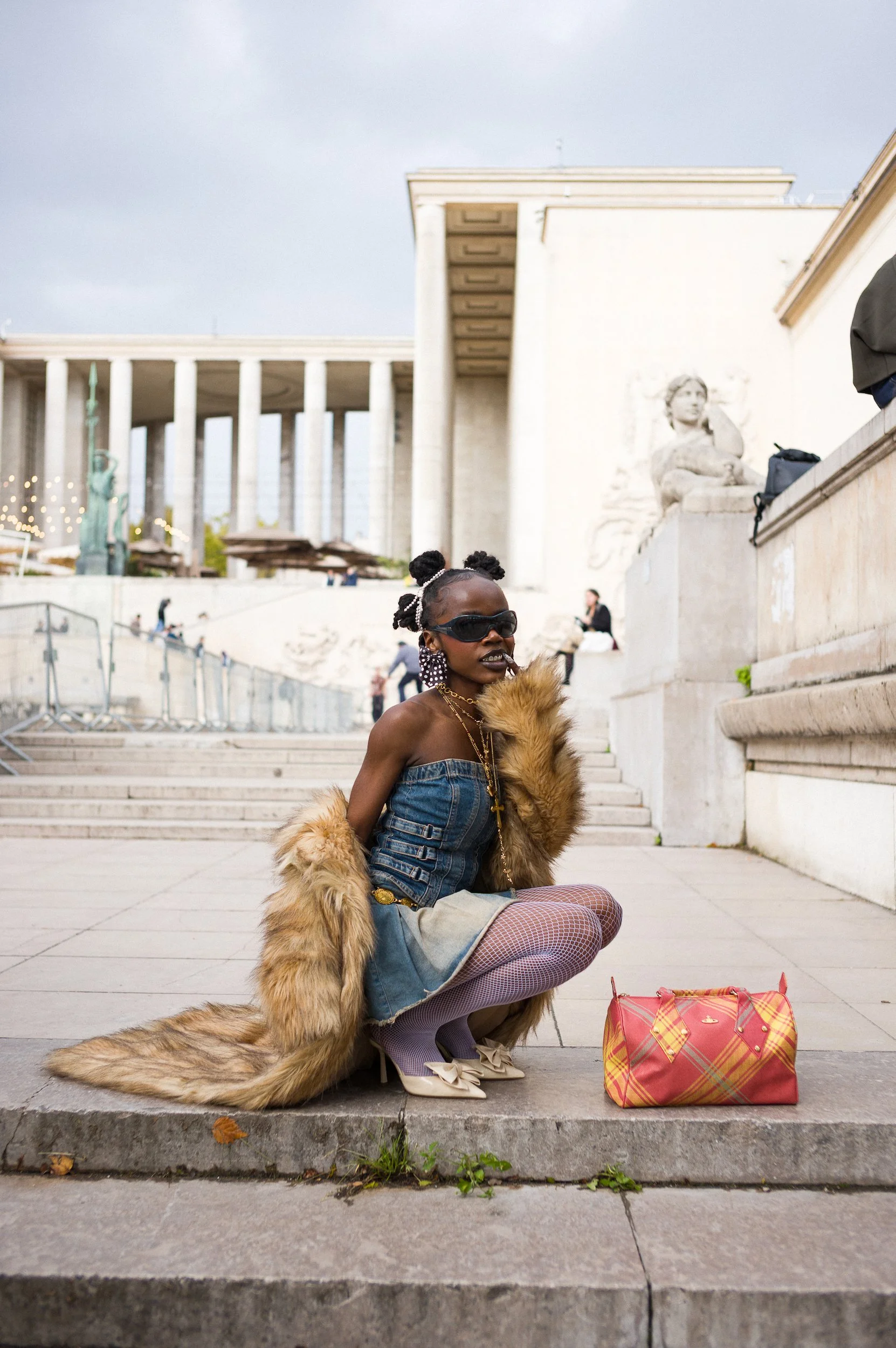 At Paris Fashion WA person dressed in a mix of fashion styles, wearing a fur coat, fishnet stockings, a denim top, and accessories, squatting on steps in front of a historic building with classical columns and sculptures, with a red plaid bag nearby.