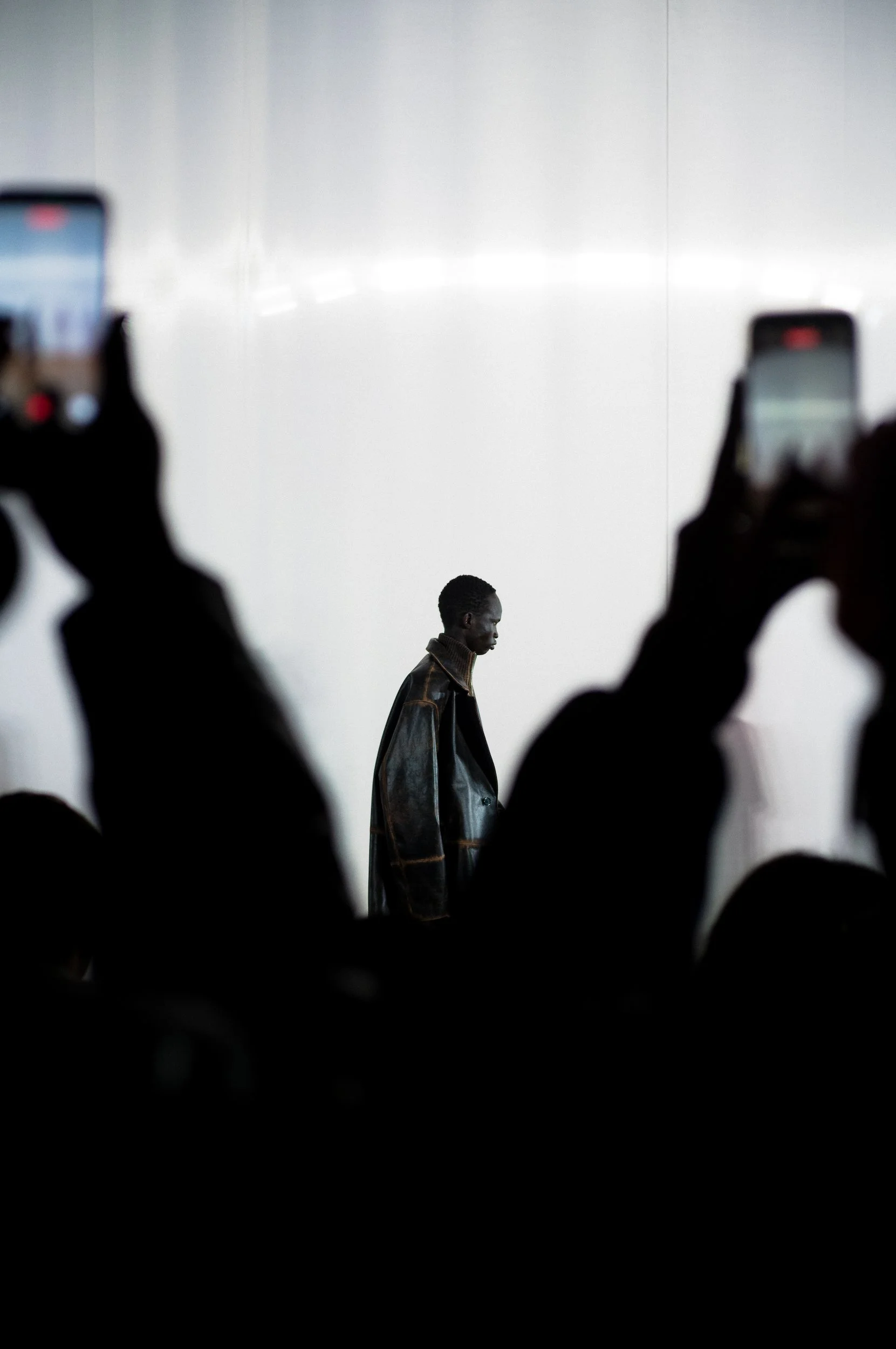 At Paris Fashion Week, A person with dark skin walking across a plain bright background, with blurred hands and phones in the foreground holding up devices to take pictures or videos.