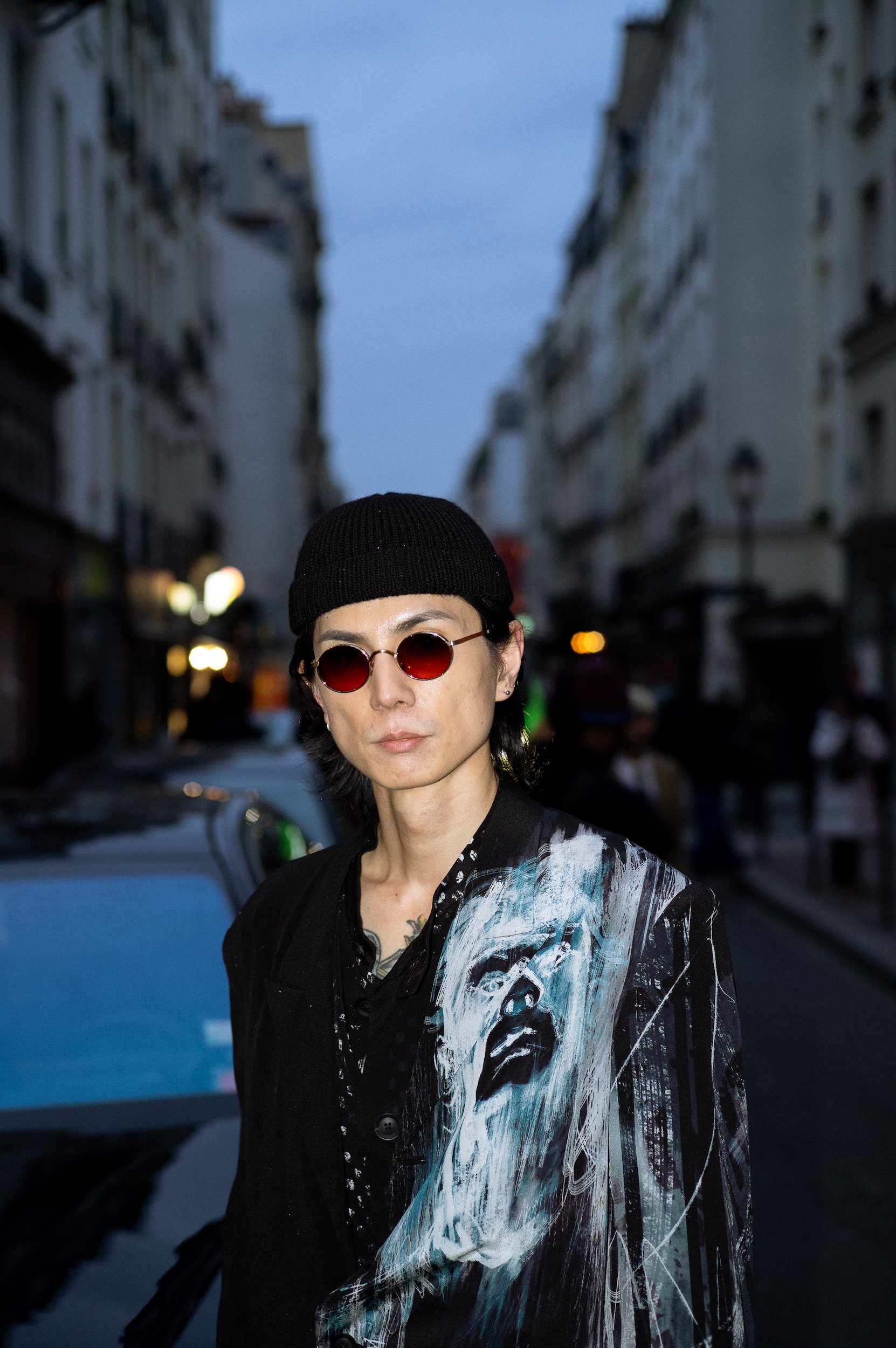 At Paris Fashion Week, Person wearing black beanie, round red sunglasses, and a black and white artistic jacket standing on a city street during dusk.