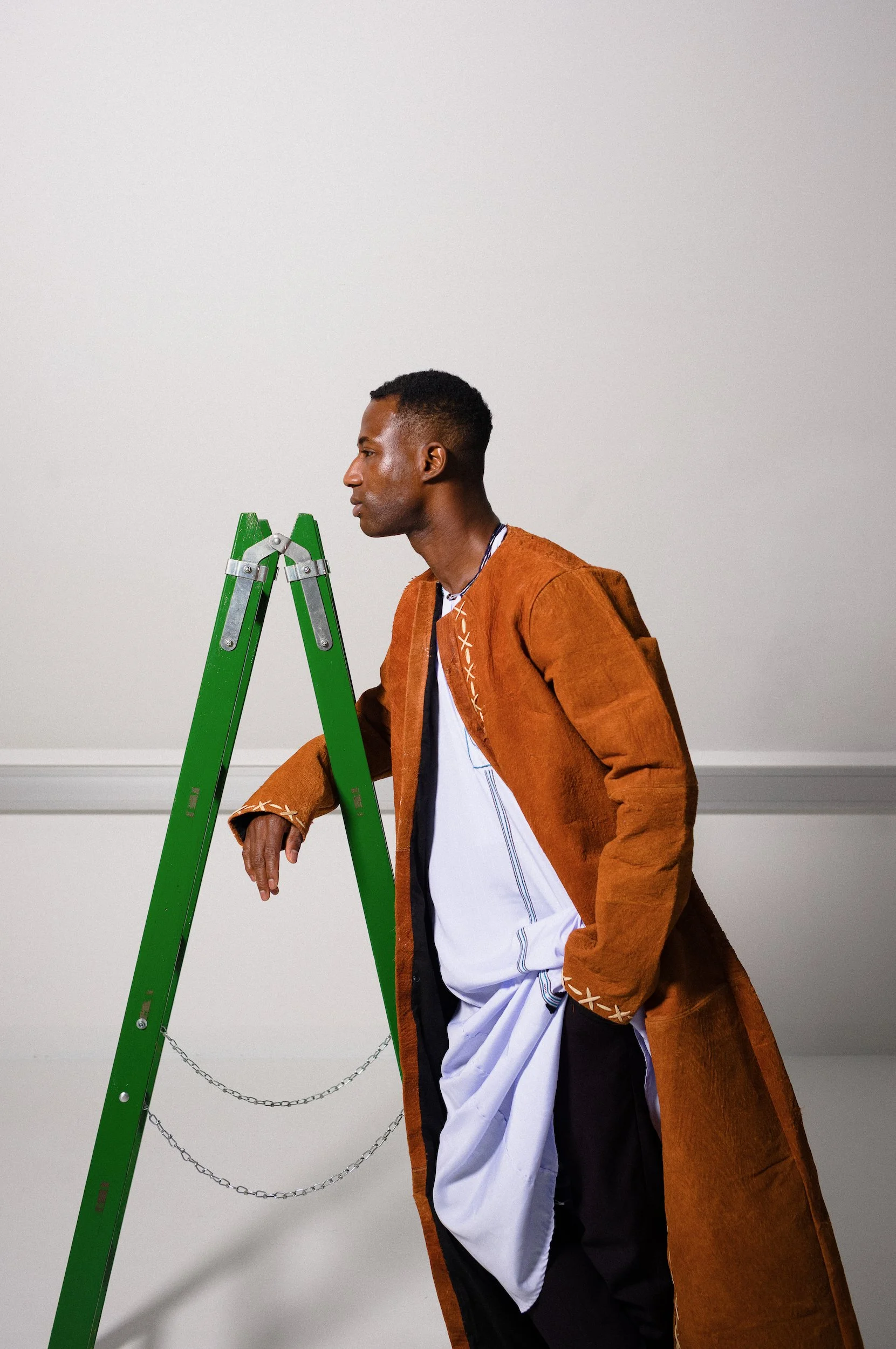 A man wearing an orange coat, black and white clothing, and a silver chain stands next to a green step ladder with chains hanging from it, positioned in front of a plain white wall.