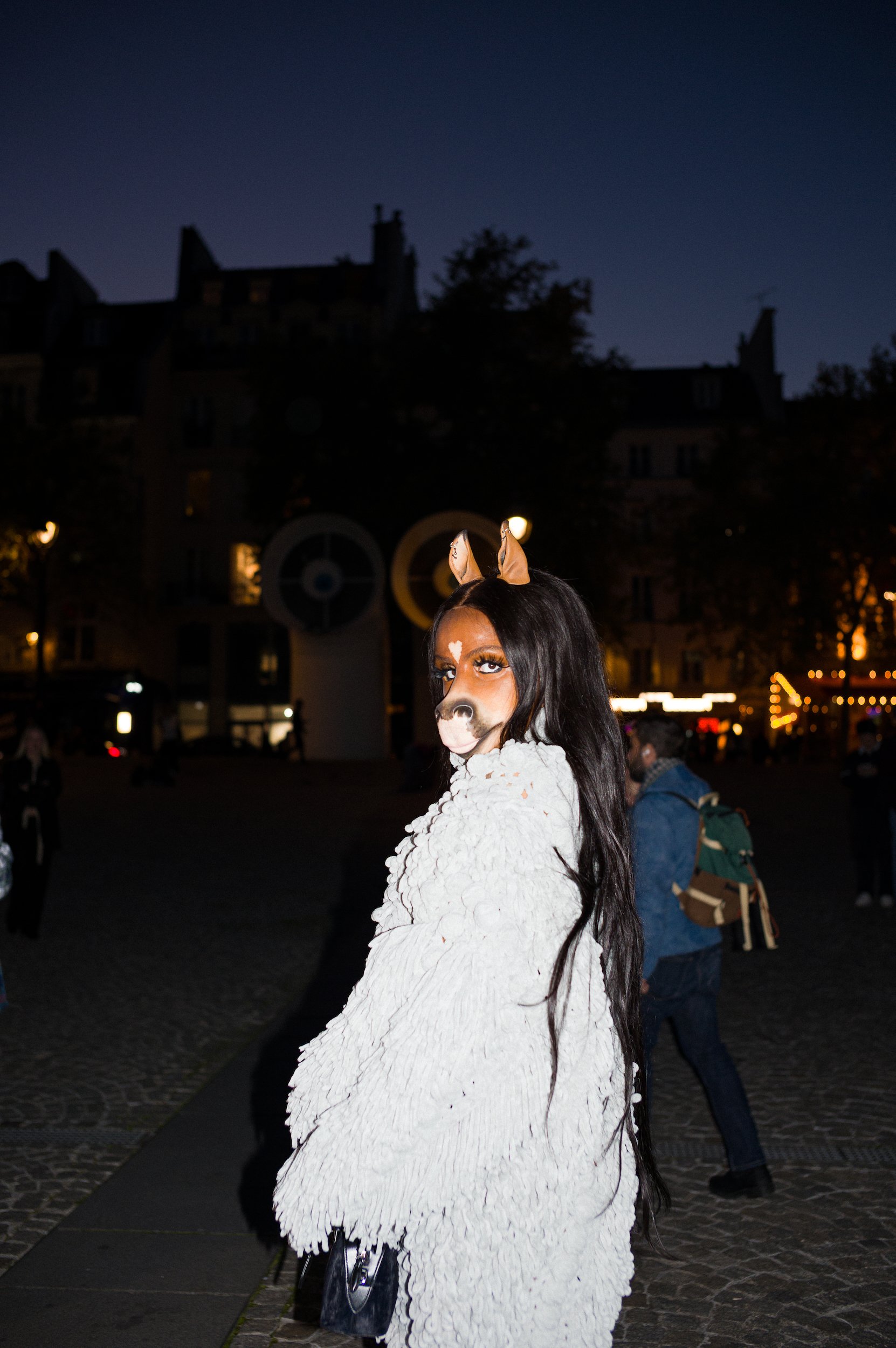A woman is dressed in a white fluffy coat with a horse face and ears painted or worn on her face, and she is standing outdoors at night in an urban area with buildings and string lights in the background.