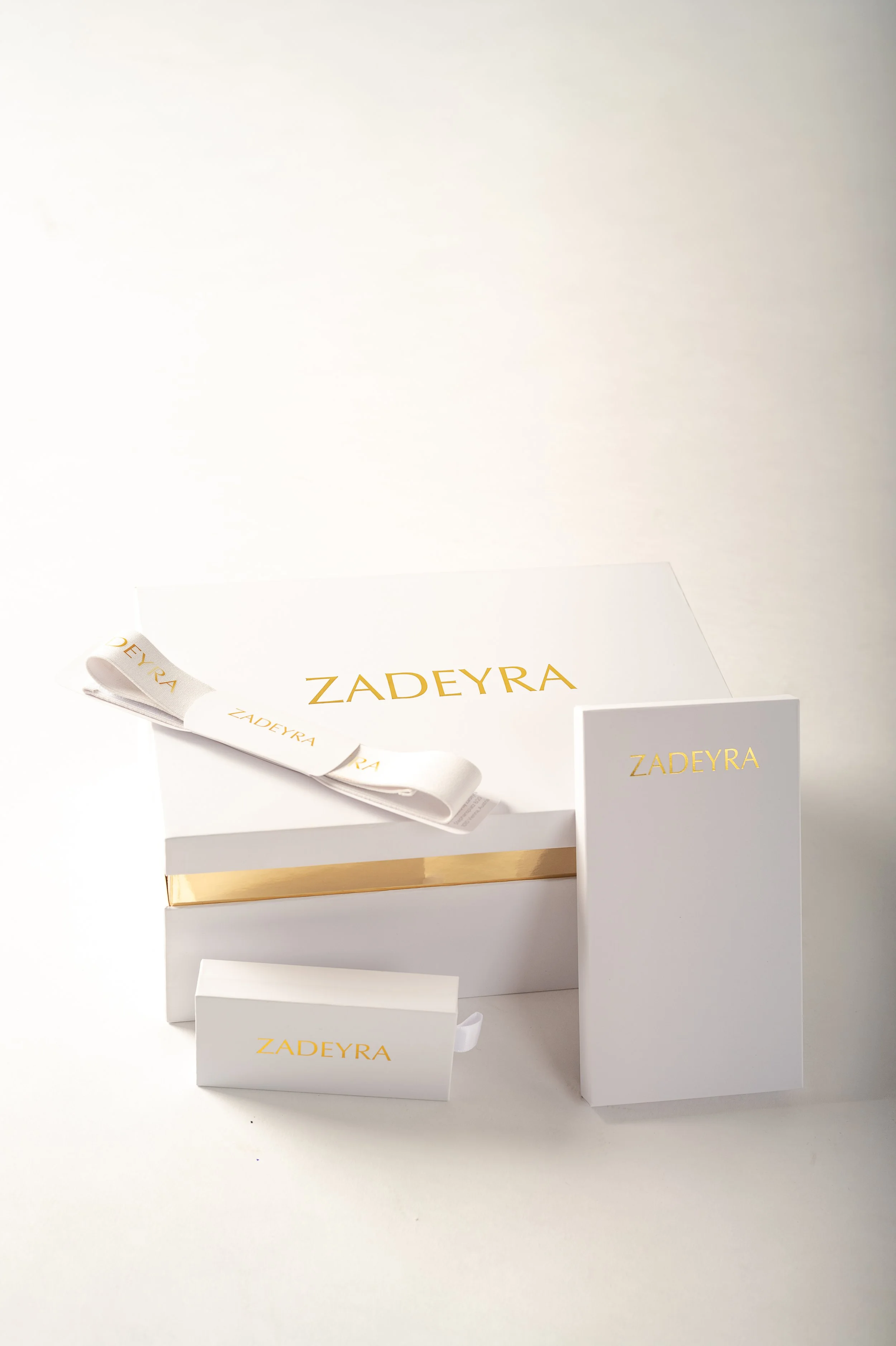 A display of white packaging boxes and a ribbon with the brand name 'ZADEYRA' in gold lettering, arranged on a white surface.