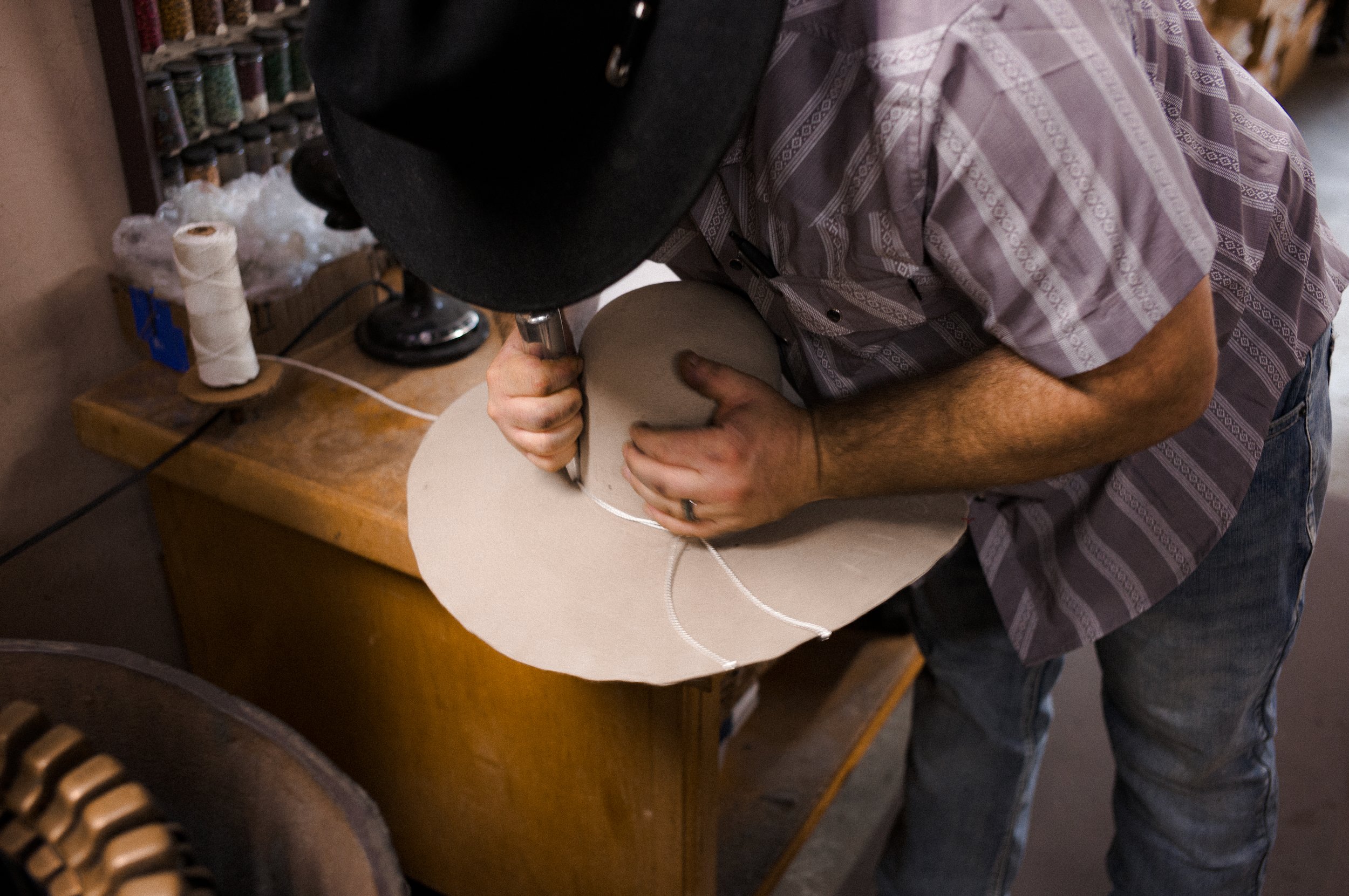 A person is shaping the crown of a cowboy hat on a workbench in a workshop. The person is wearing a plaid shirt and a black cap.