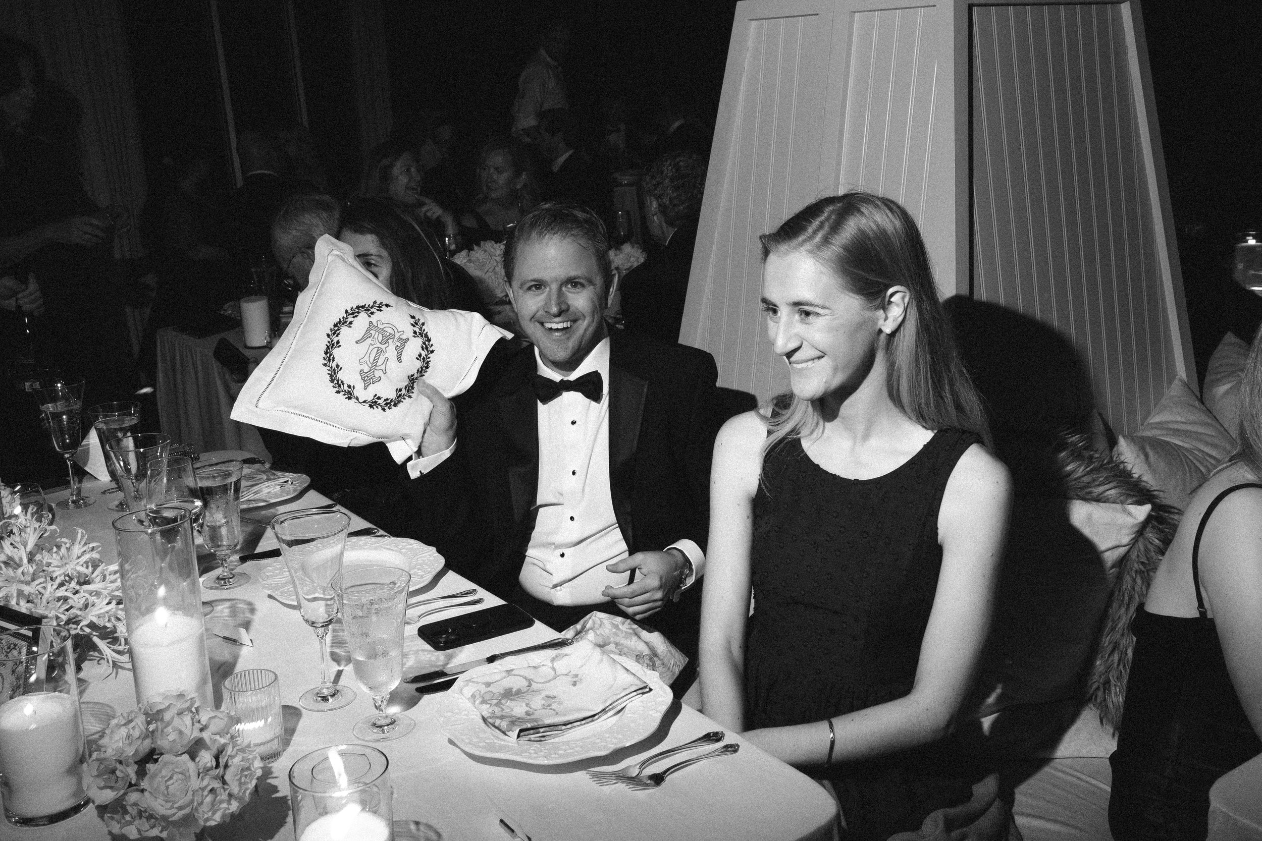 A black and white photo of a man in a tuxedo holding a pillow with an insignia, sitting next to a woman in a dress at a formal dinner event. The table is set with glassware, plates, and floral decorations, with other guests visible in the background.