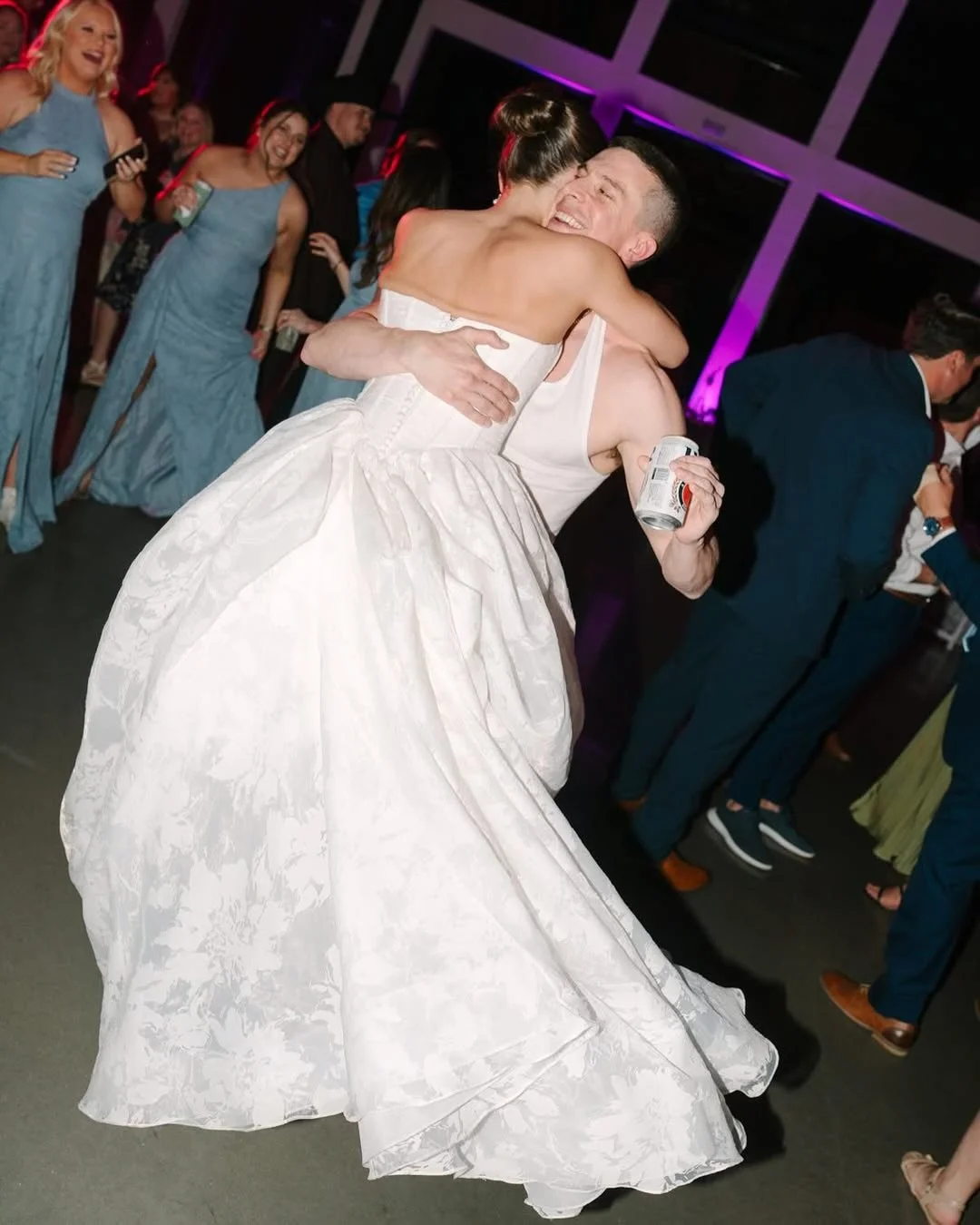 A woman in a wedding dress and a man in a sleeveless shirt hugging and celebrating at a wedding reception, with guests dancing and laughing in the background.