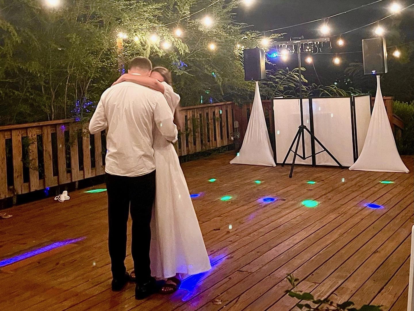 A couple dancing on a wooden deck at night with string lights overhead, a DJ setup with speakers and lighting in the background, surrounded by trees.