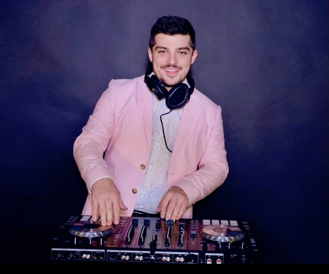 A young man with dark hair and a beard, wearing a pink blazer and headphones around his neck, standing behind a DJ console with a dark background.