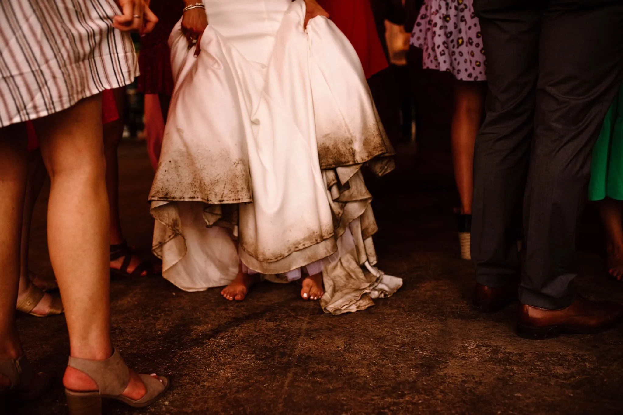 A group of people dressed in formal attire, standing close together in a dimly lit indoor setting. The focus is on the lower half of their bodies, with a woman in a white gown, likely a wedding dress, in the center. The dress has dirt stains on the bottom, and her toes are visible. Surrounding her are other individuals wearing heels, dress shoes, and skirts or pants.