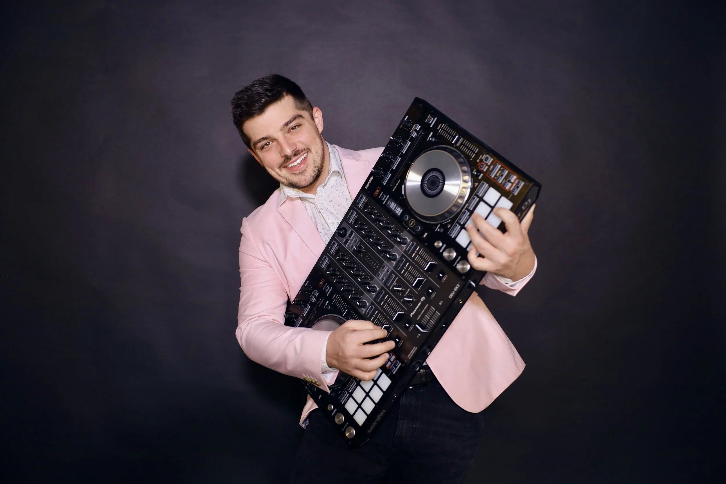 A man smiling and holding a DJ controller against a dark background, wearing a light pink blazer and a white shirt.
