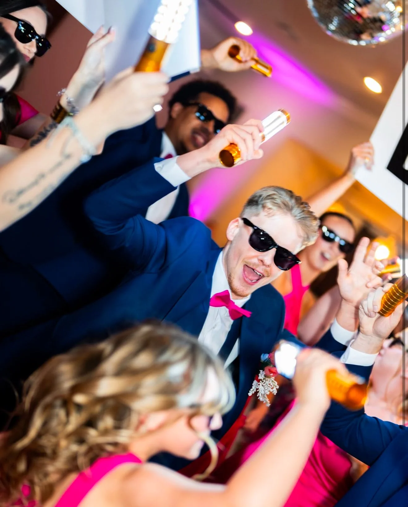 People celebrating at a party with drinks, wearing sunglasses, and dressed up in formal attire with a festive background.