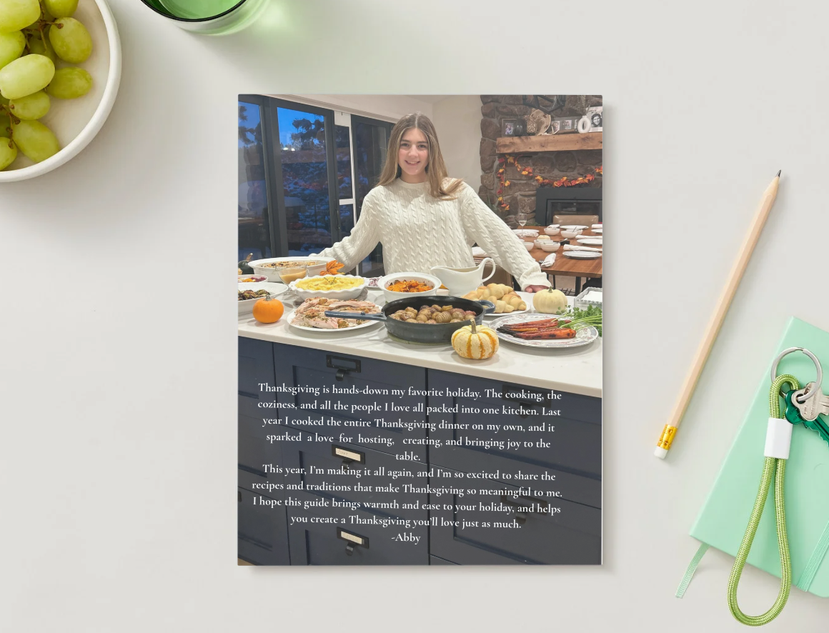 A printed Thanksgiving greeting card resting on a light-colored surface with a bowl of green grapes, a manicured green pen, and a bundle of keys on a green notepad nearby. The card features a woman in a cozy knit sweater standing behind a table filled with Thanksgiving dishes in a warm, inviting kitchen with a stone fireplace and large windows.