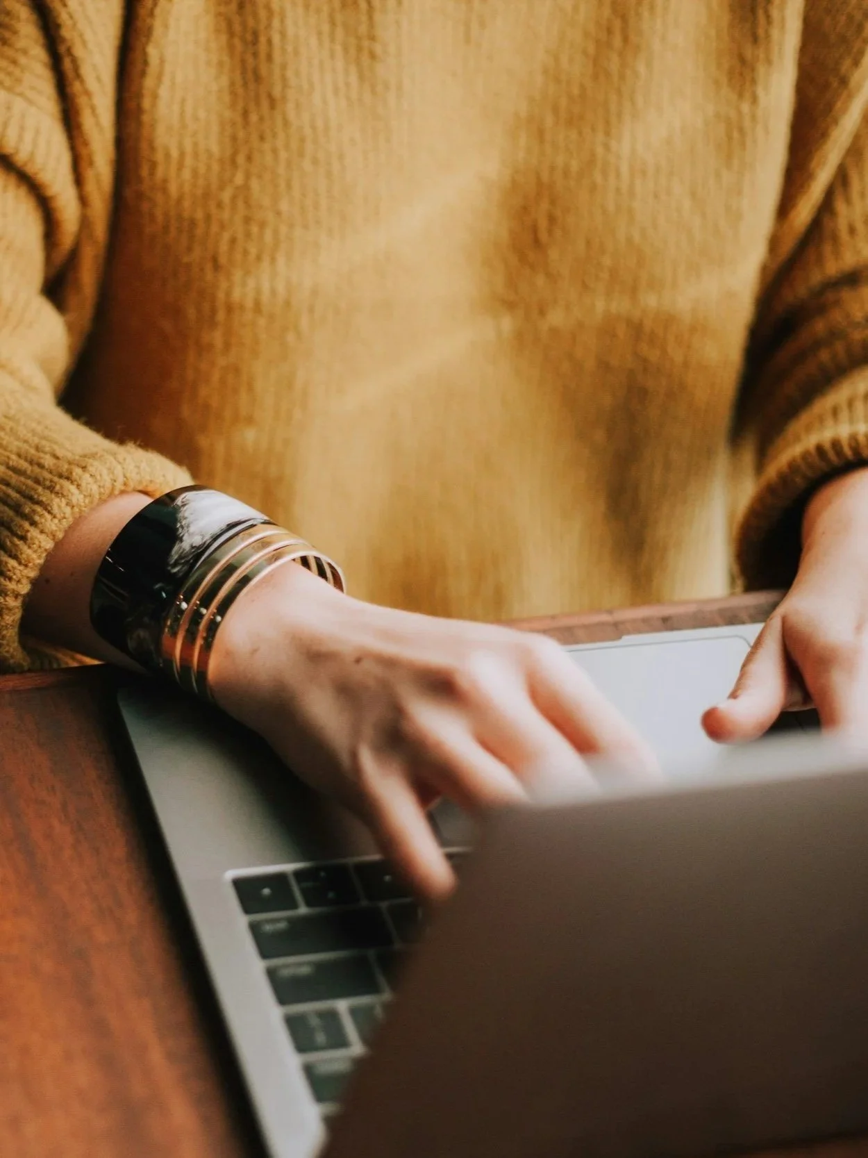 Person wearing a mustard yellow sweater with metallic bangles on the wrist, using a laptop on a wooden table.