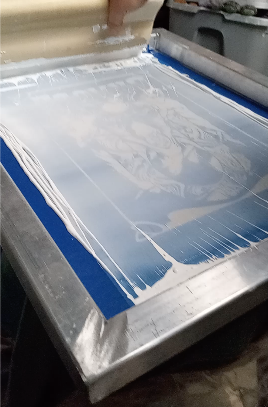 A person is pouring white liquid over a large blue screen used in the screen printing process.
