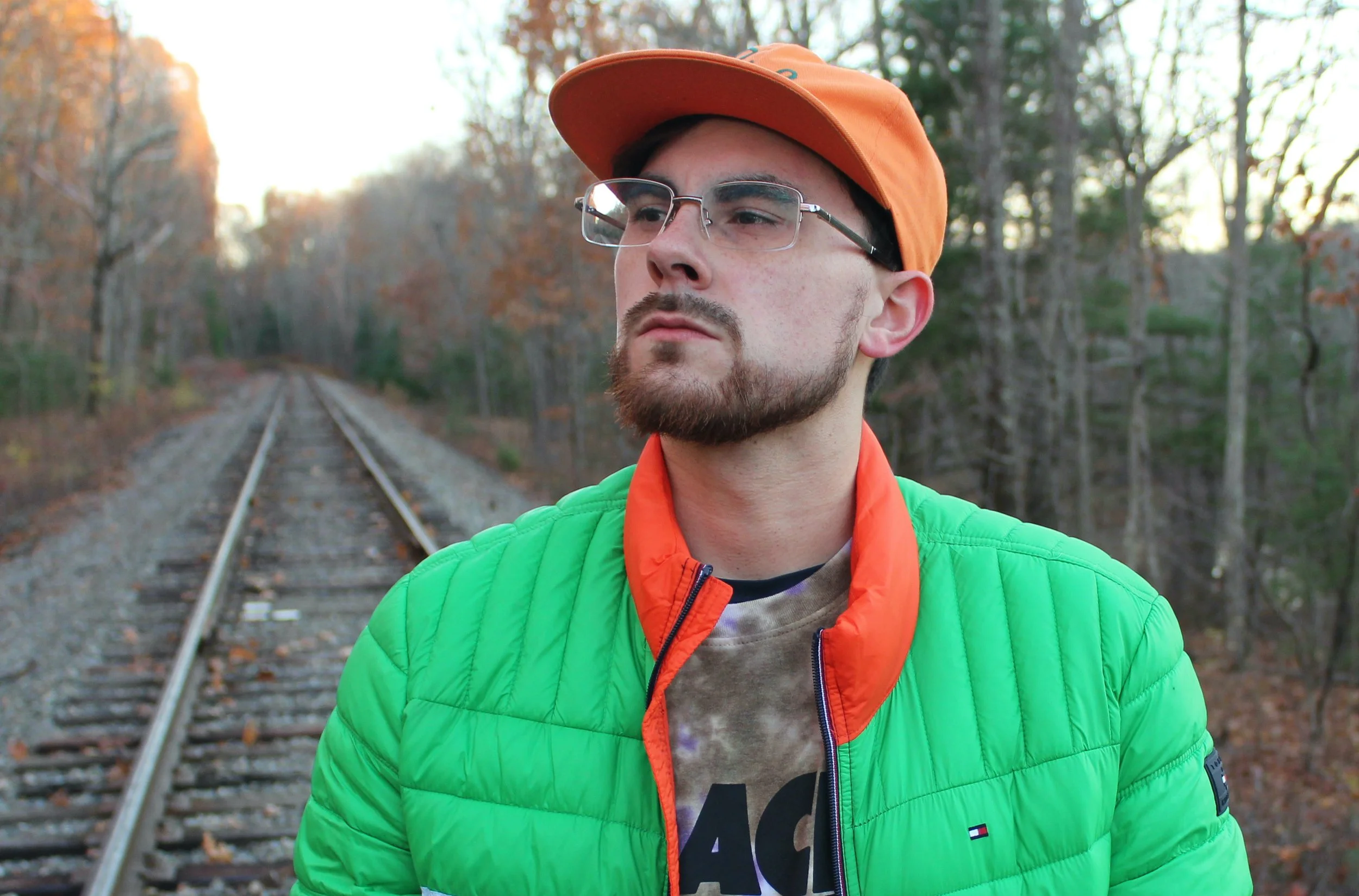 A young man with glasses, a beard, and mustache wears an orange cap and green jacket, standing on a train track in a forest with autumn leaves.