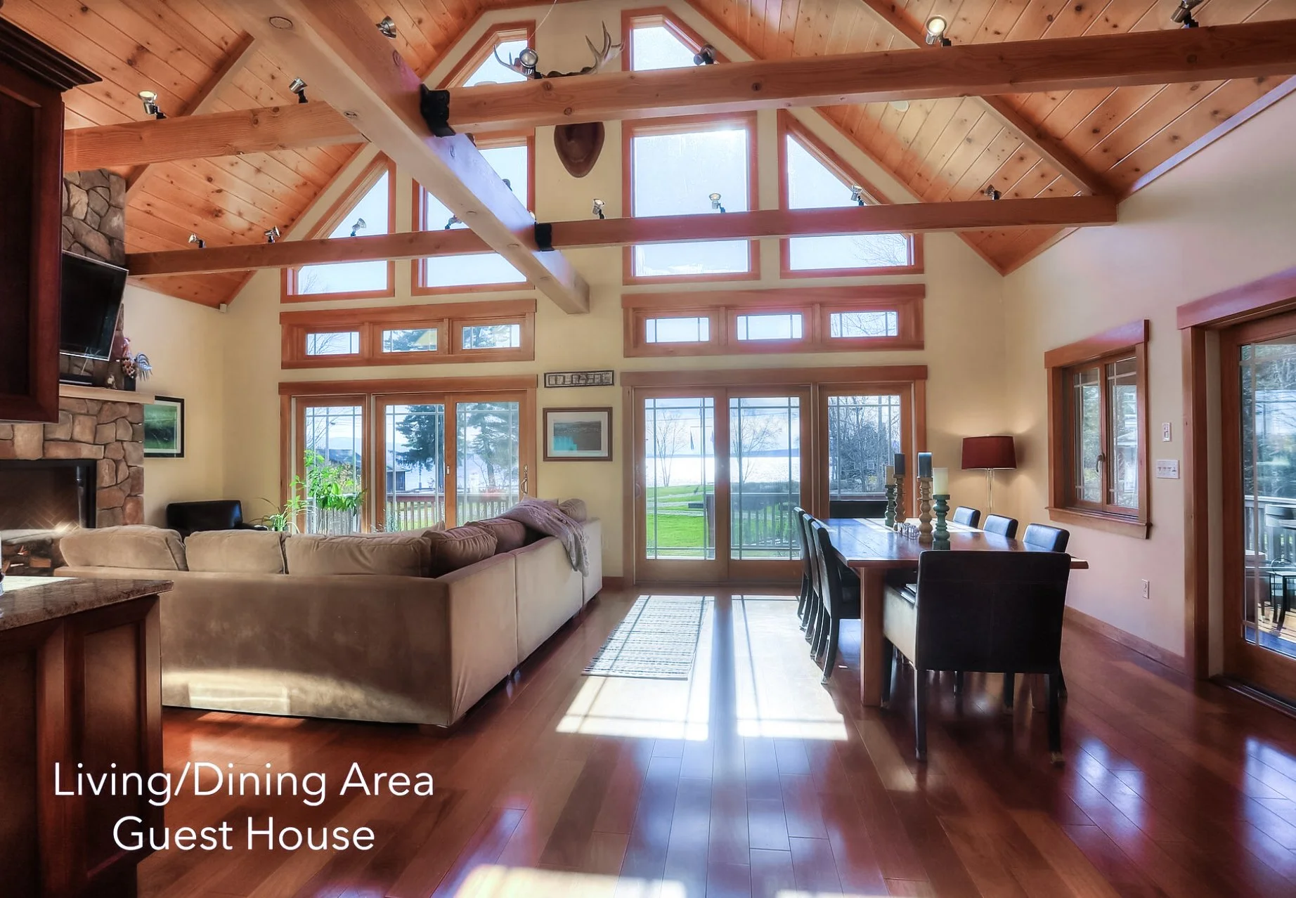 Interior of a living/dining area in a guest house with a vaulted ceiling, wooden beams, large windows and glass doors, a beige sectional sofa, a wooden dining table with black chairs, and a view of a grassy yard with lake and trees outside.