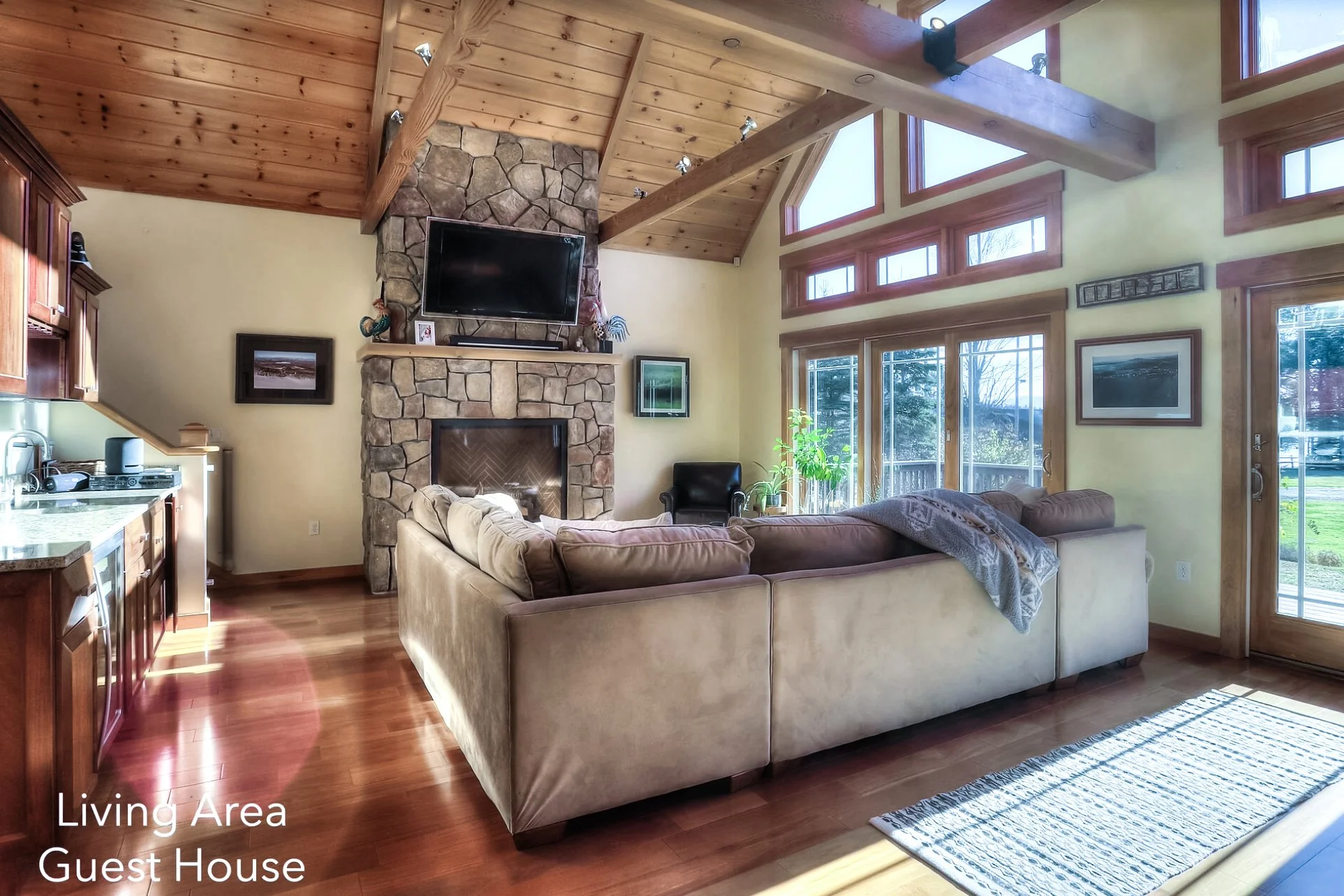 Living room in a guest house with wooden ceiling, stone fireplace, wall-mounted TV, beige sectional sofa, black armchair, large windows, glass sliding door, hardwood floor, potted plants, and framed pictures.