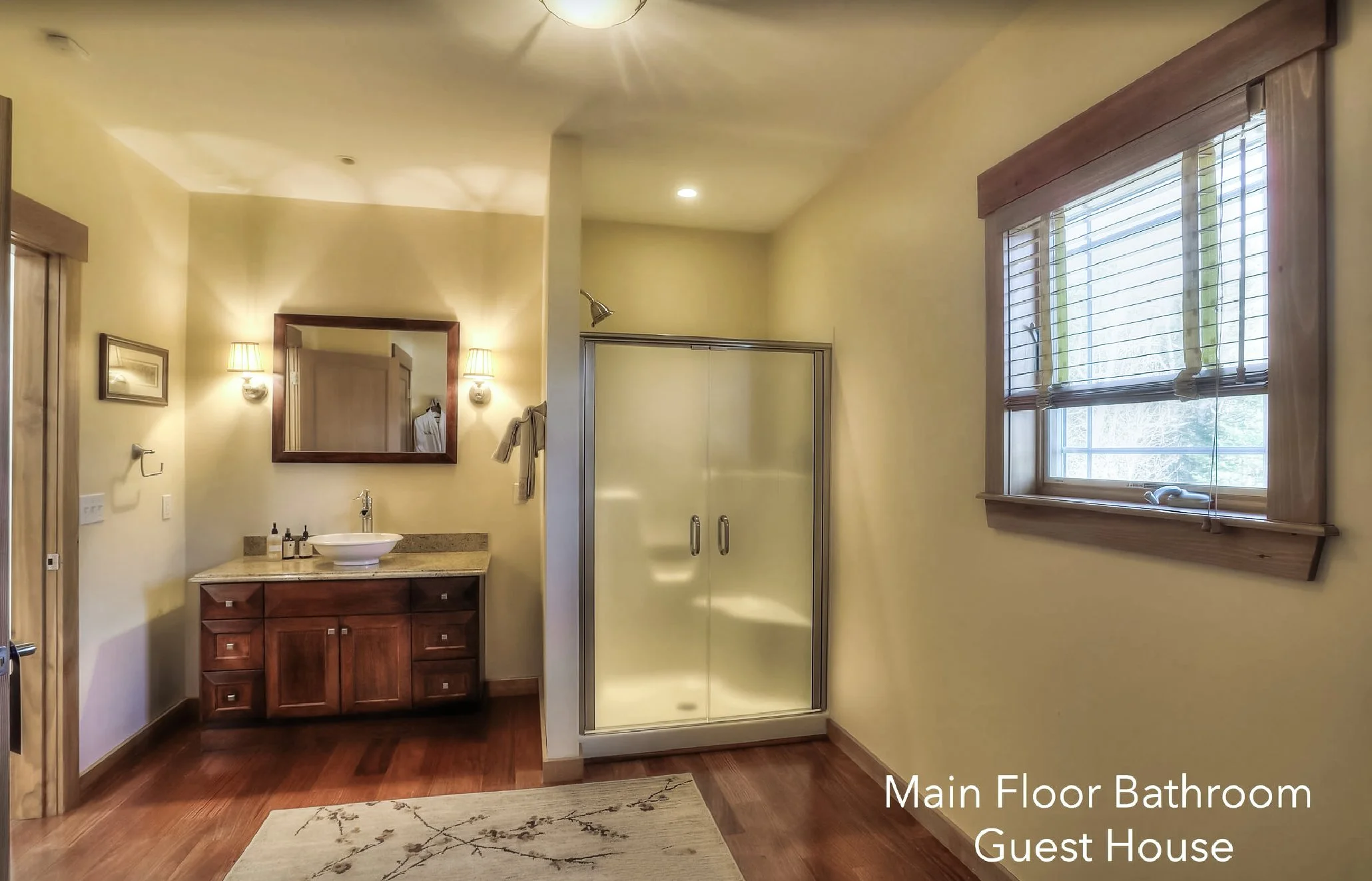 Bathroom with wooden vanity, white vessel sink, mirror, wall-mounted lights, shower stall with glass door, window with wooden frame and blinds, beige walls, hardwood floor, and a rug. Text overlay says 'Main Floor Bathroom Guest House'.