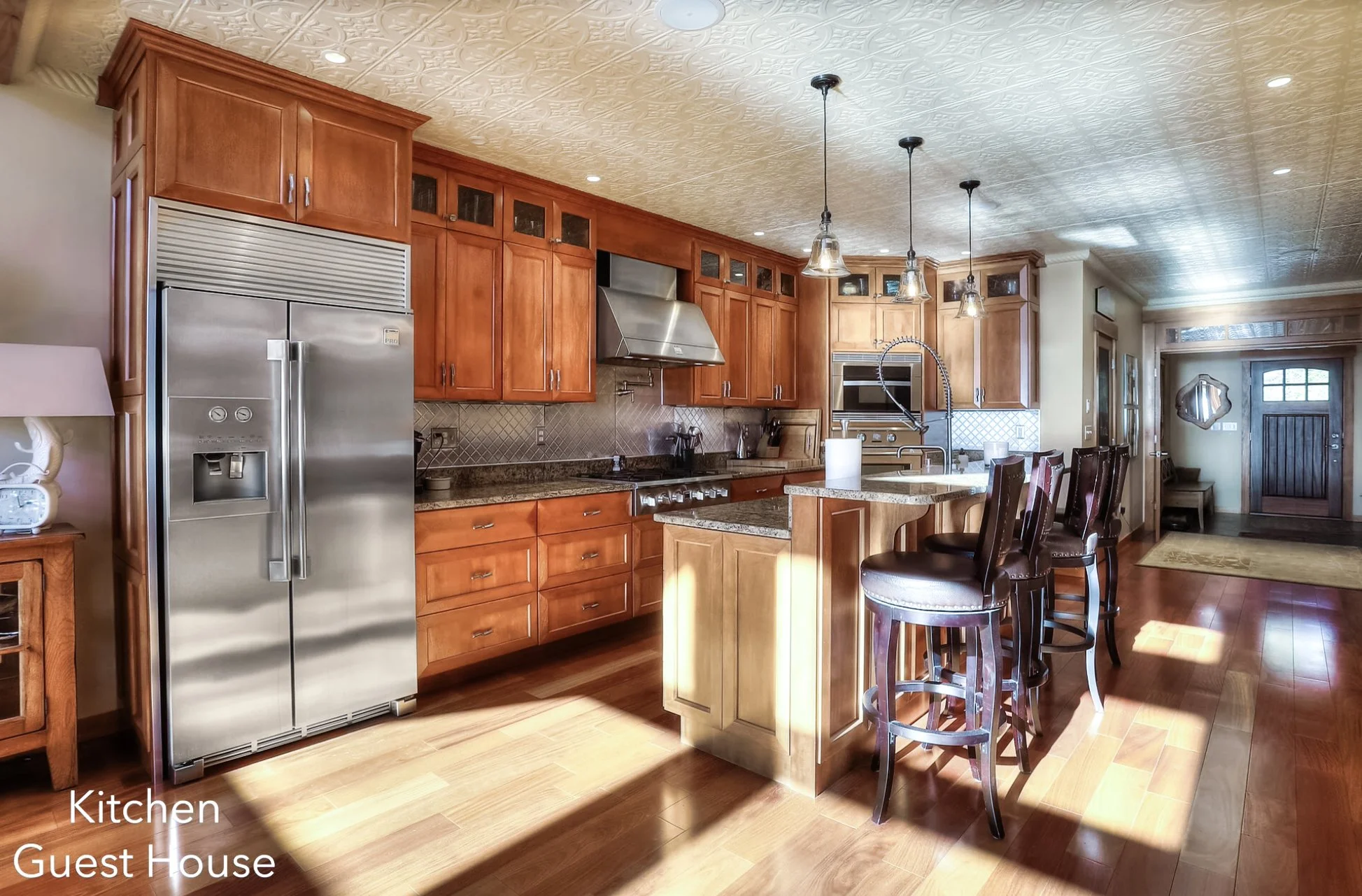 Kitchen in a guest house with wooden cabinetry, stainless steel refrigerator, granite countertops, hanging pendant lights, and a kitchen island with bar stools.