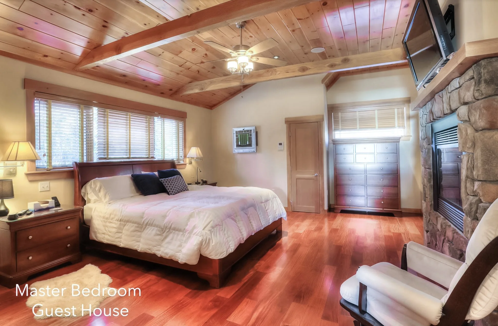 A spacious master bedroom with a large bed, wooden furniture, a stone fireplace, and a ceiling fan, featuring hardwood floors and multiple windows with blinds.