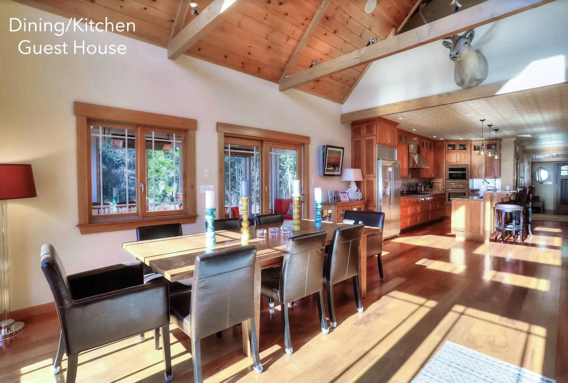 Open-concept dining and kitchen area in a guest house with wooden beams, hardwood floors, large windows, and a mounted deer head. The dining table has six chairs and colorful candleholders, and the kitchen features wooden cabinets and a breakfast bar