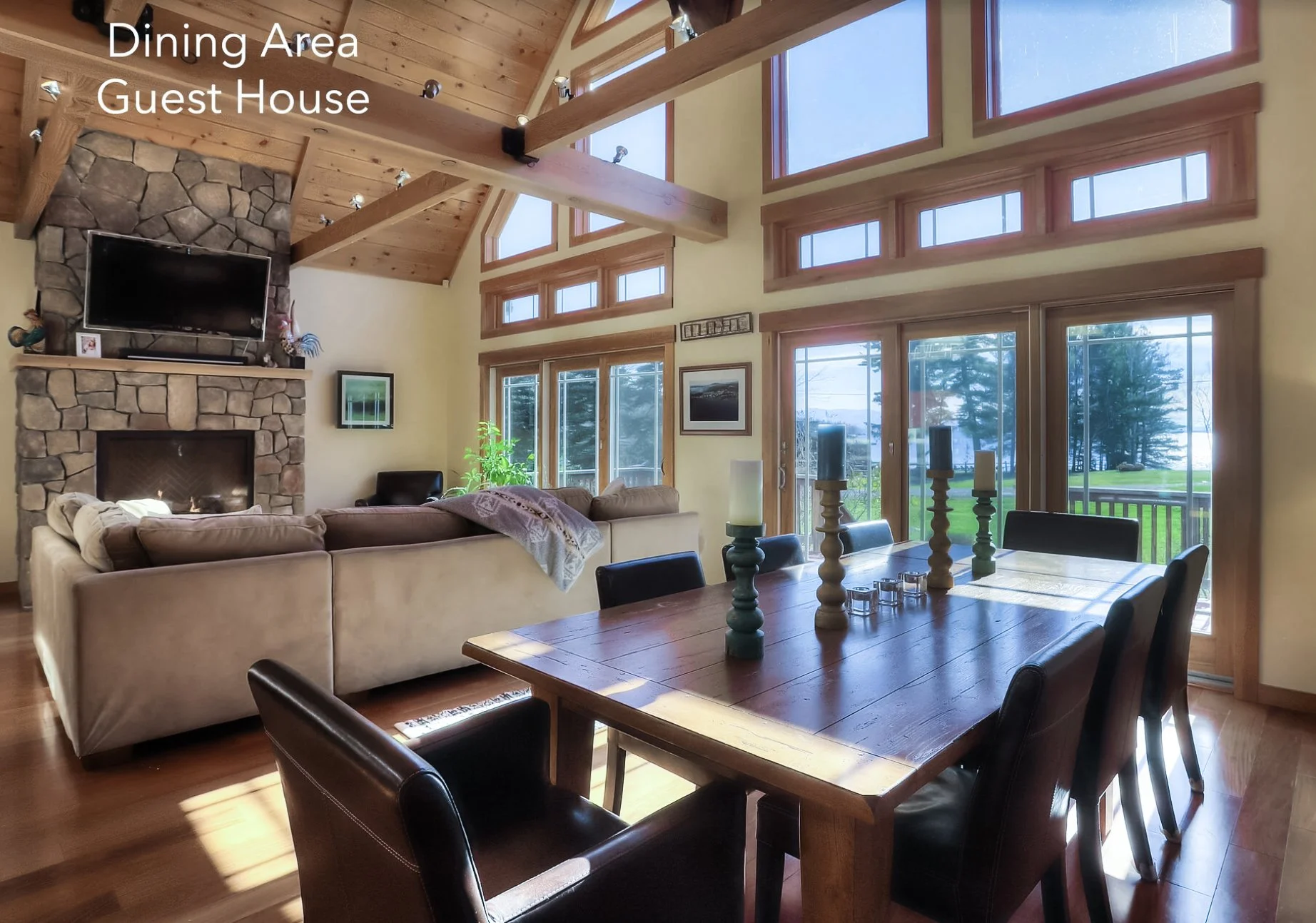 A living room with a stone fireplace, a TV above it, a large beige sectional sofa, a wooden dining table with candles, large windows showing a green outdoor landscape, and a vaulted wooden ceiling with exposed beams.