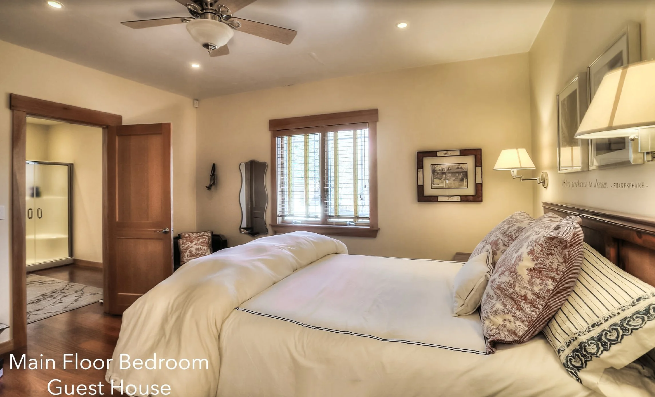 Main floor bedroom in guest house with bed, pillows, window with blinds, wall art, and wall-mounted lamps