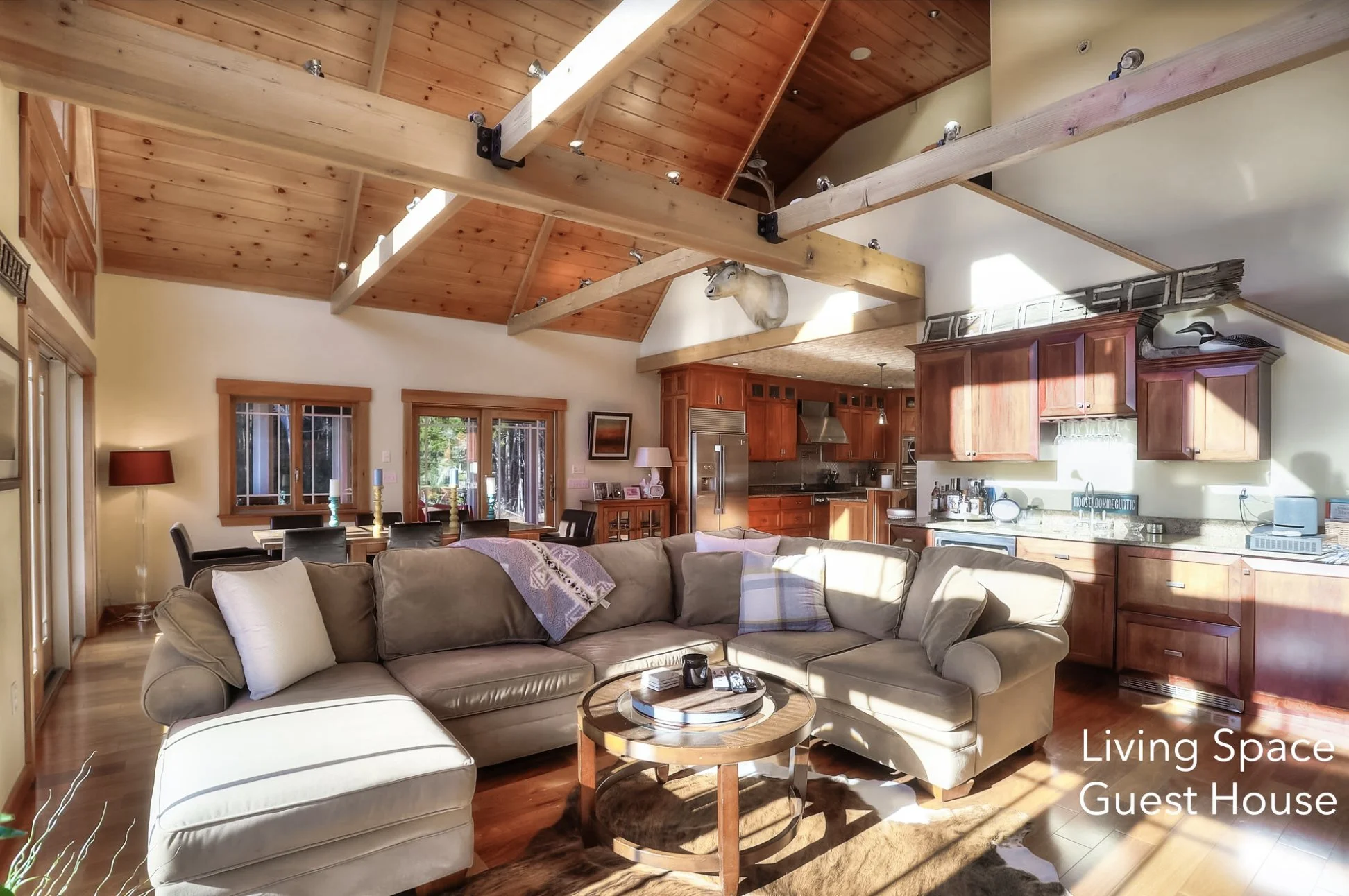 Interior of a living space guest house with a beige sectional sofa, wooden coffee table, kitchen area with wooden cabinets, and large windows showing outdoor scenery. Exposed wooden beams on the ceiling, and natural sunlight illuminating the room.