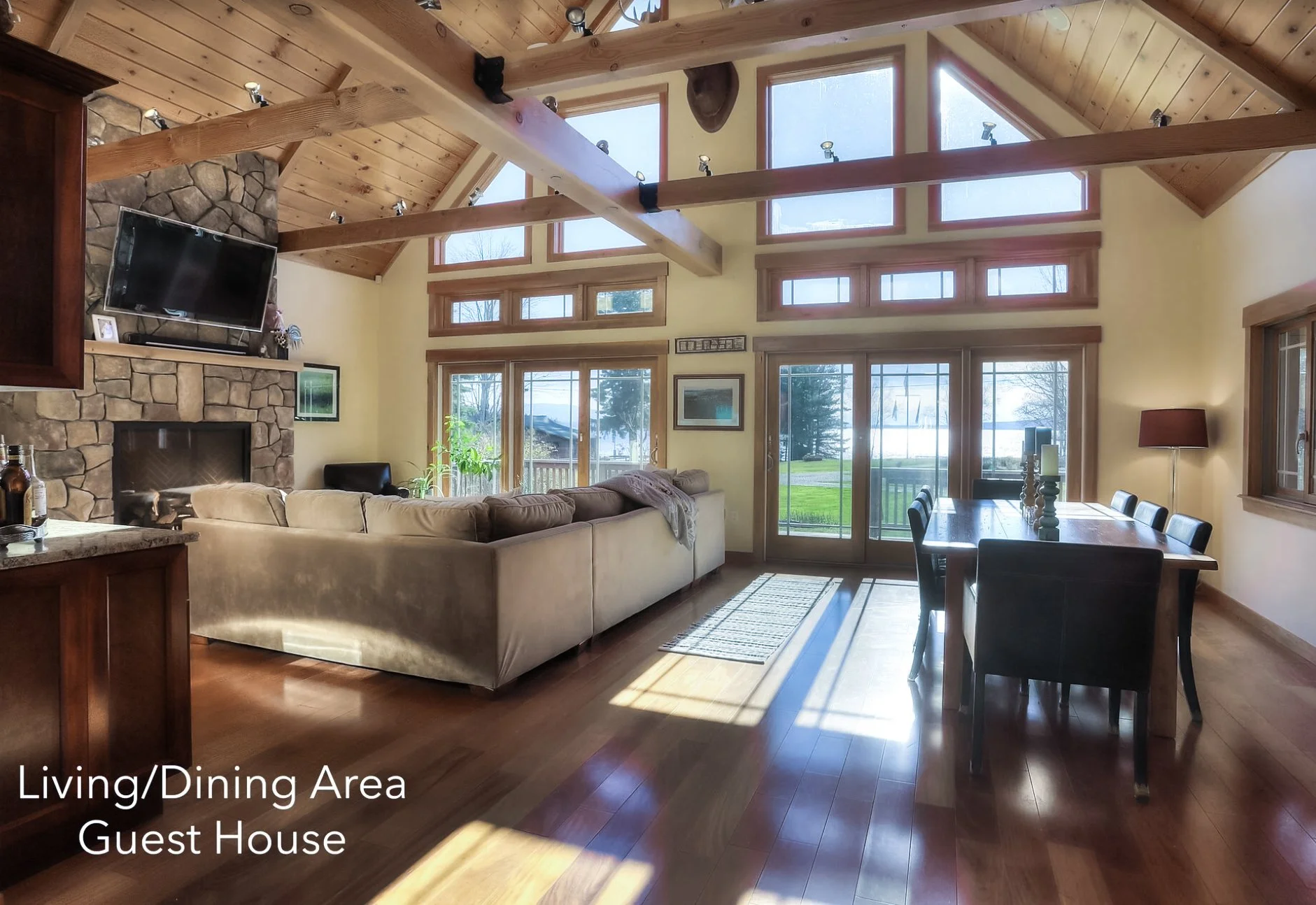 Interior of a living/dining area in a guest house with a high, peaked wooden ceiling. Features include a stone fireplace with a mounted TV, a large beige sectional sofa, a rectangular dining table with black chairs, and large windows and glass doors 