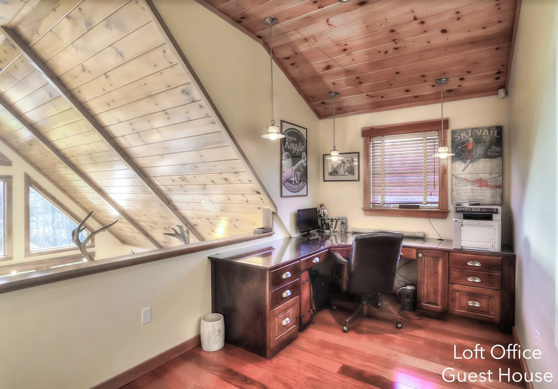 Loft office in a guest house with a wooden desk, black office chair, computer, printer, and wall decor, featuring wooden ceiling and flooring, and a small window with blinds.