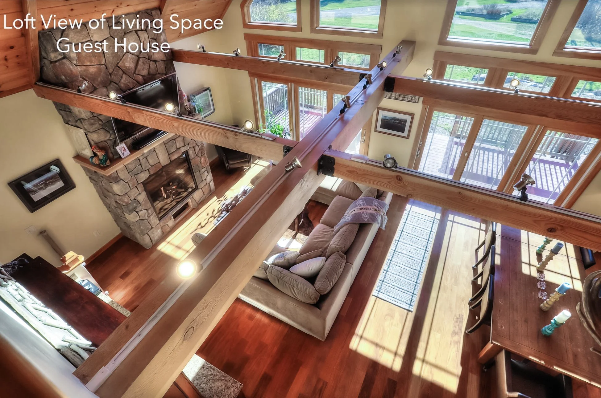 Loft view of living space in a guest house showing a cozy living room with a stone fireplace, wooden beams, large windows, a beige sofa with pillows, a wooden dining table with colorful candles, and a sliding glass door leading to a deck with outdoor