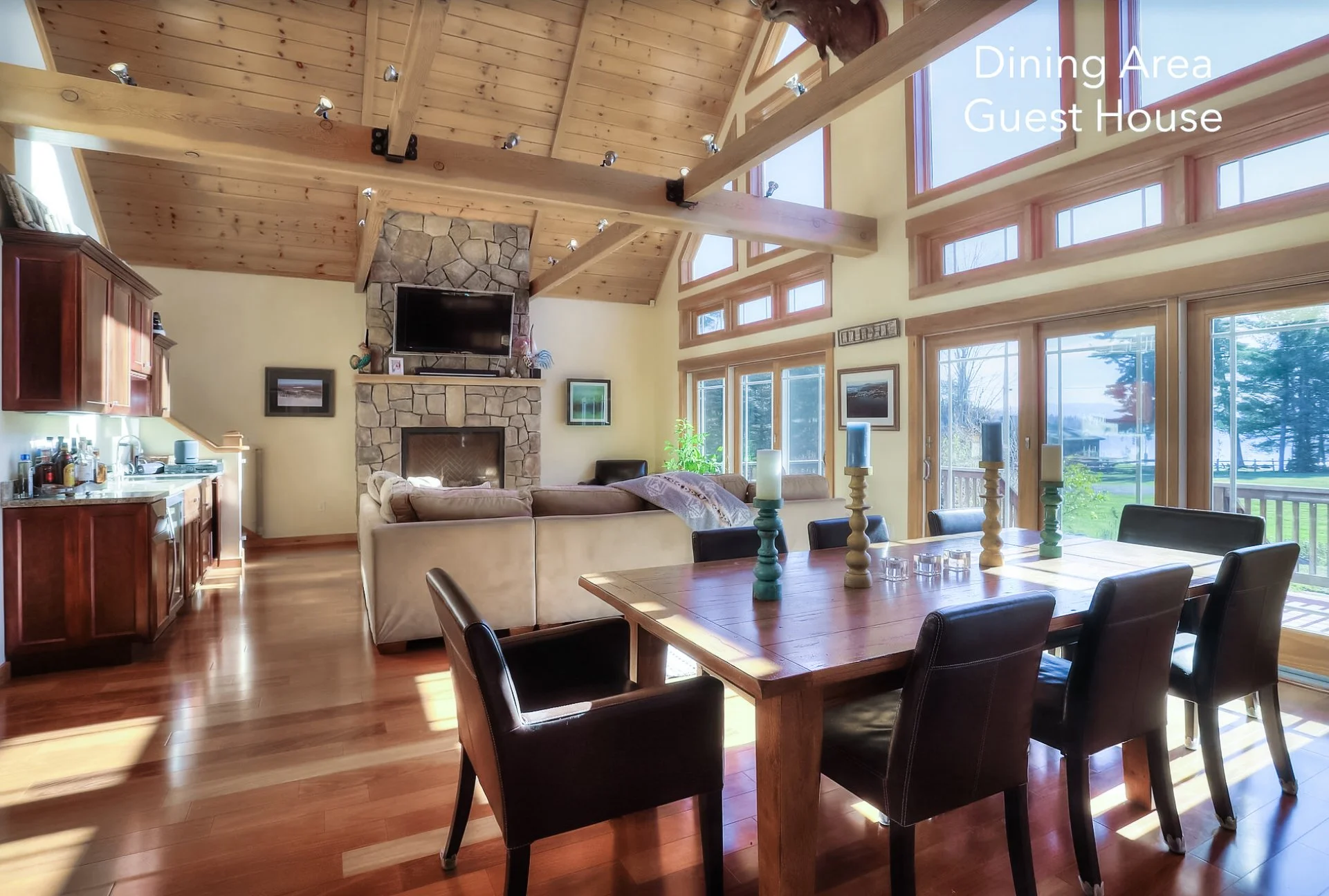 Interior of a dining area and guest house with wooden ceiling, large windows, and a couch facing a stone fireplace with a TV mounted above.