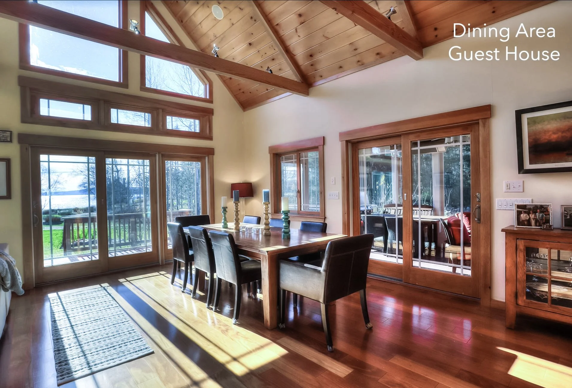 Interior view of a dining area guest house with wooden ceiling beams, large windows, a sliding glass door leading outside, a wooden dining table with six black chairs, and candles on the table. Text in the top right corner reads 'Dining Area Guest Ho