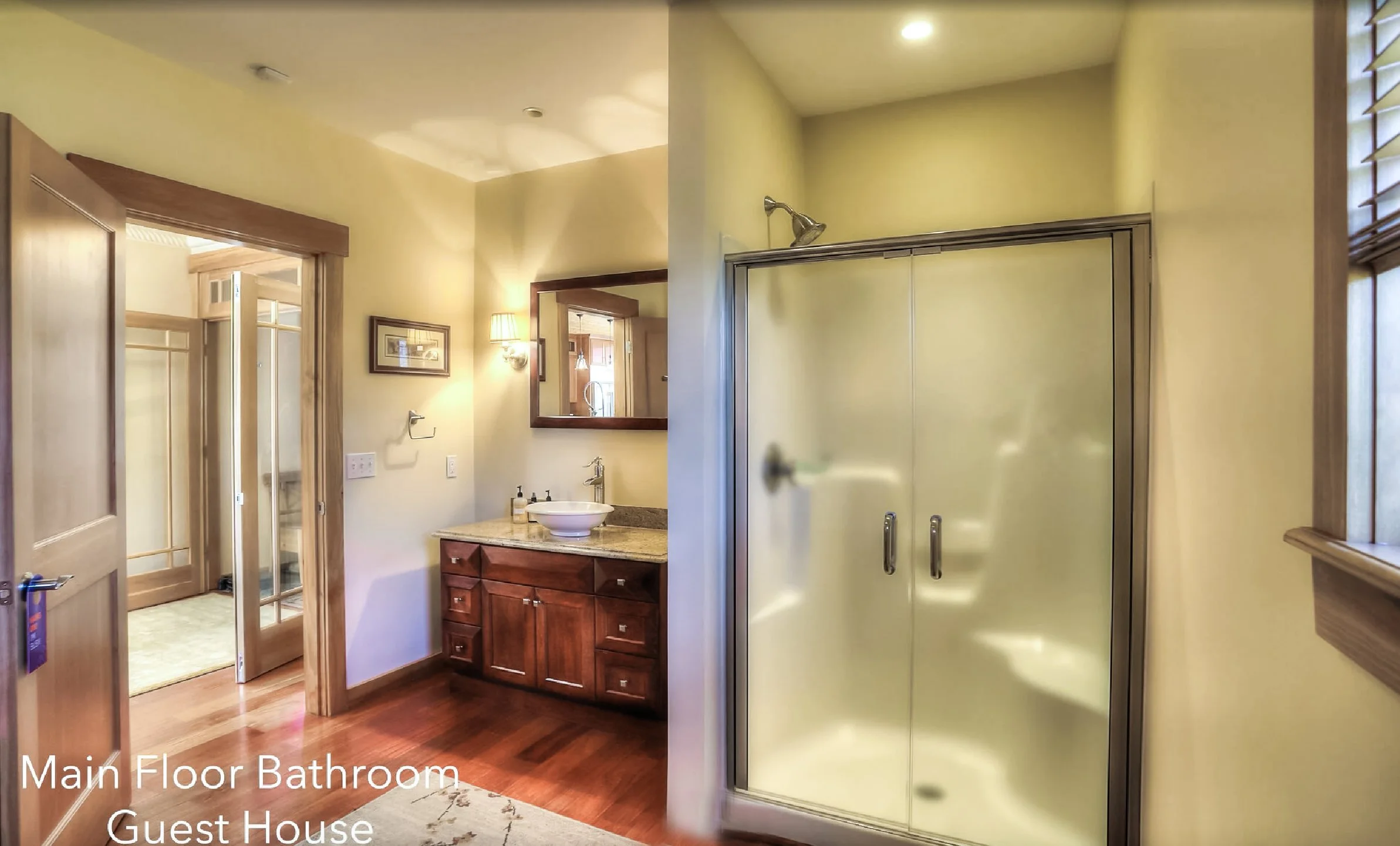 Main floor bathroom in a guest house with a wooden vanity, a sink, a mirror, a shower with frosted glass doors, a window with blinds, and framed artwork on the wall.