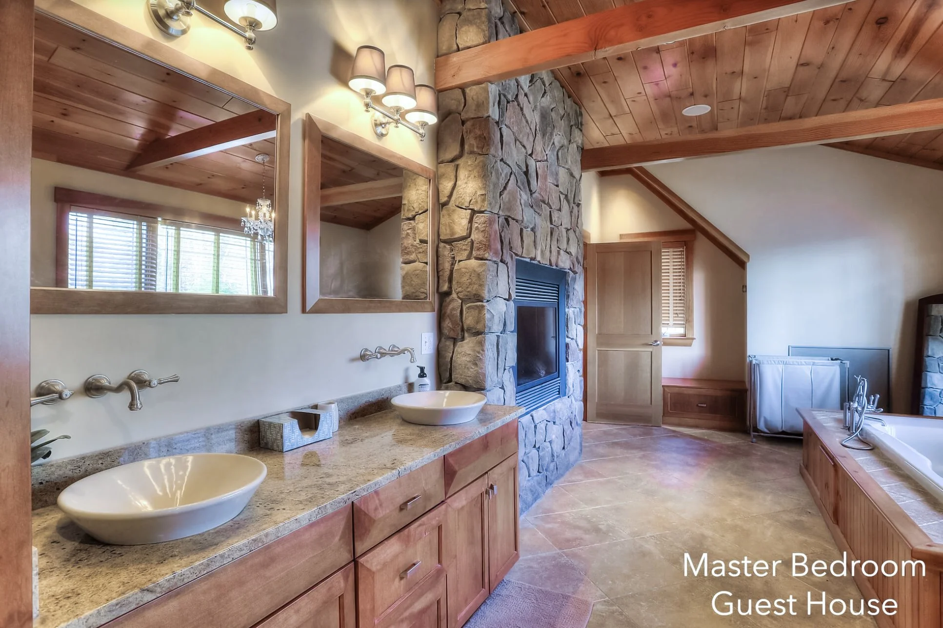 A spacious master bathroom in a guest house with a double sink vanity, large mirrors, stone accents around the fireplace, wooden countertops, and a bathtub. The bathroom features wooden ceiling beams and a mix of stone and wood decor.