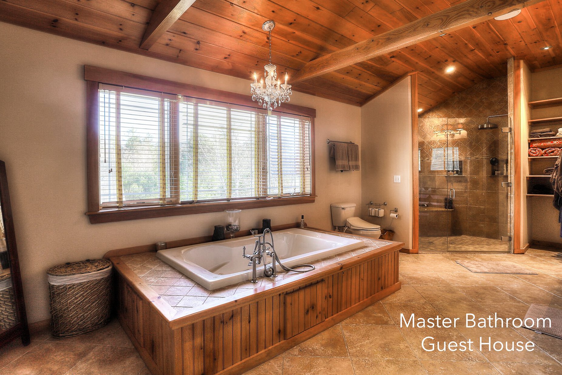 A spacious master bathroom with a wooden ceiling, large window with wooden blinds, a chandelier, a built-in bathtub with tile surround, a glass-enclosed shower, and built-in shelves with towels, labeled 'Master Bathroom Guest House'.