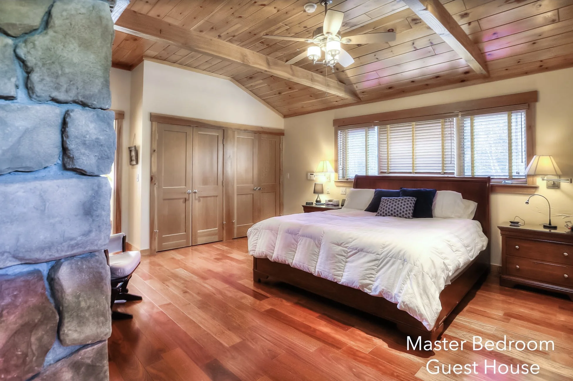 A master bedroom in a guest house with wood-paneled vaulted ceiling, hardwood flooring, a large bed with white bedding and black and gray pillows, two bedside tables with lamps, and a large window with wooden blinds.