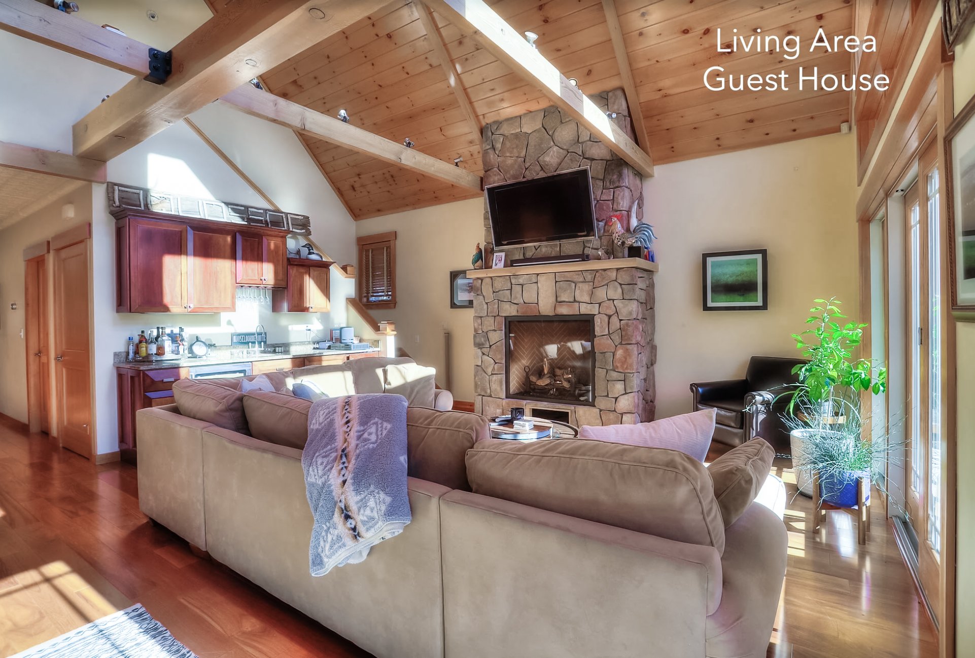 Living area with beige sofa, black chair, stone fireplace with mounted TV, wood ceiling beams, and a large window with a potted plant nearby.