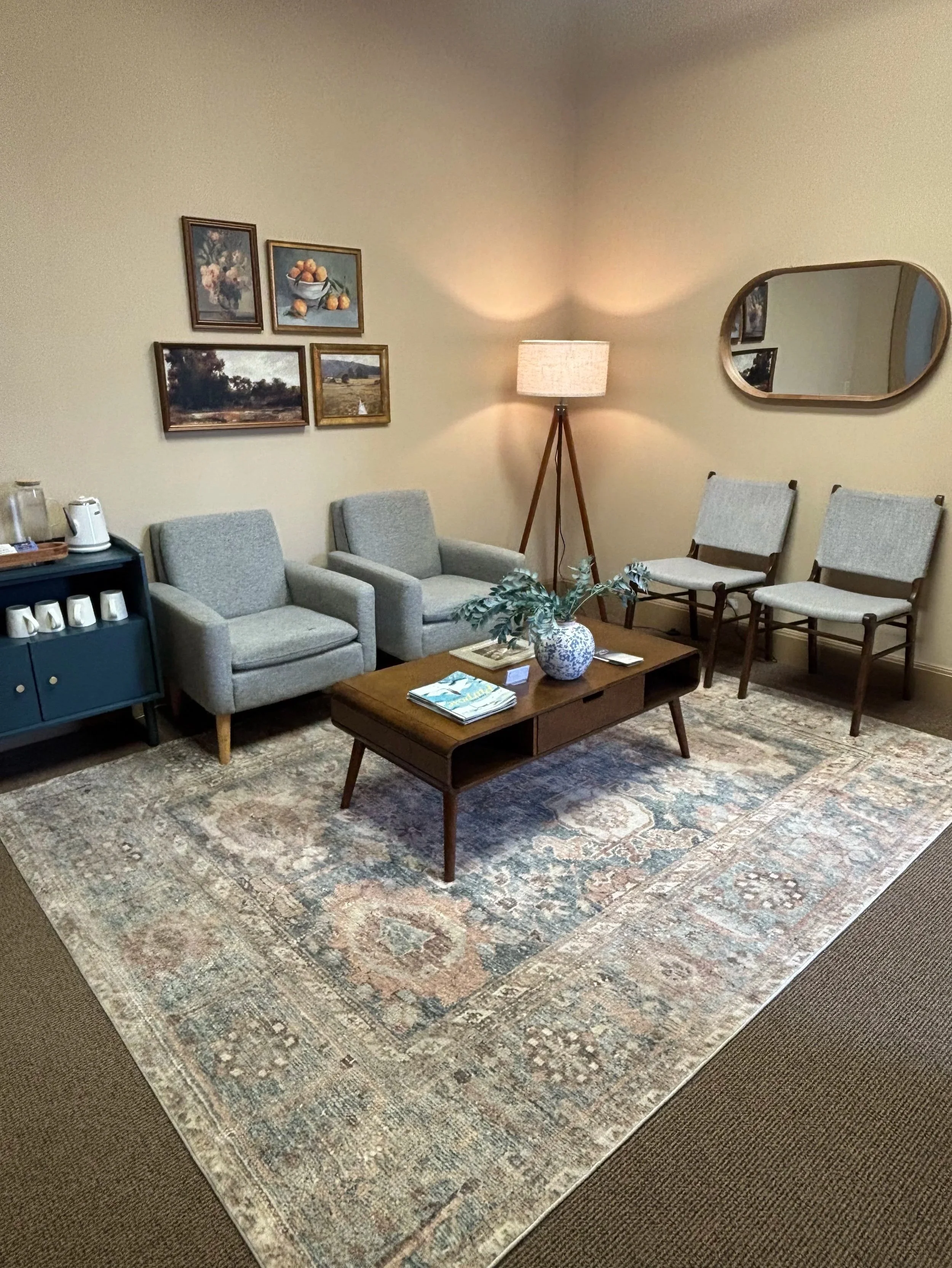 A waiting room with a blue and beige rug, a mid century modern coffee table, and four gray chairs for clients to sit in. There is a mirror on one wall and a gallery wall of art prints on the other. There is a table with coffee mugs and a tea kettle.